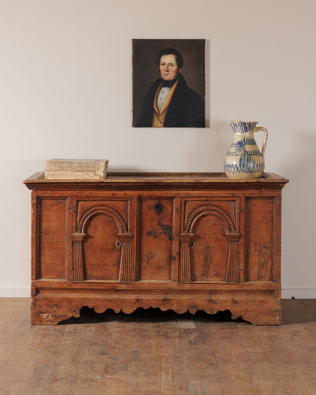 An antique carved wooden chest topped with a stack of books and a blue and white pitcher, beneath a portrait of a man.