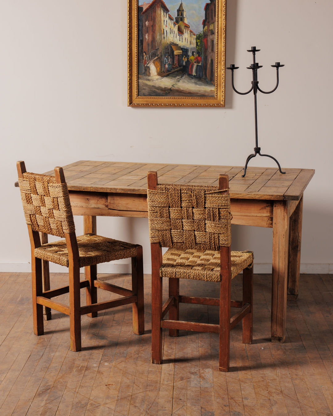 A rustic wooden table with two woven-back chairs, a framed painting of a European street, and an iron candelabra.
