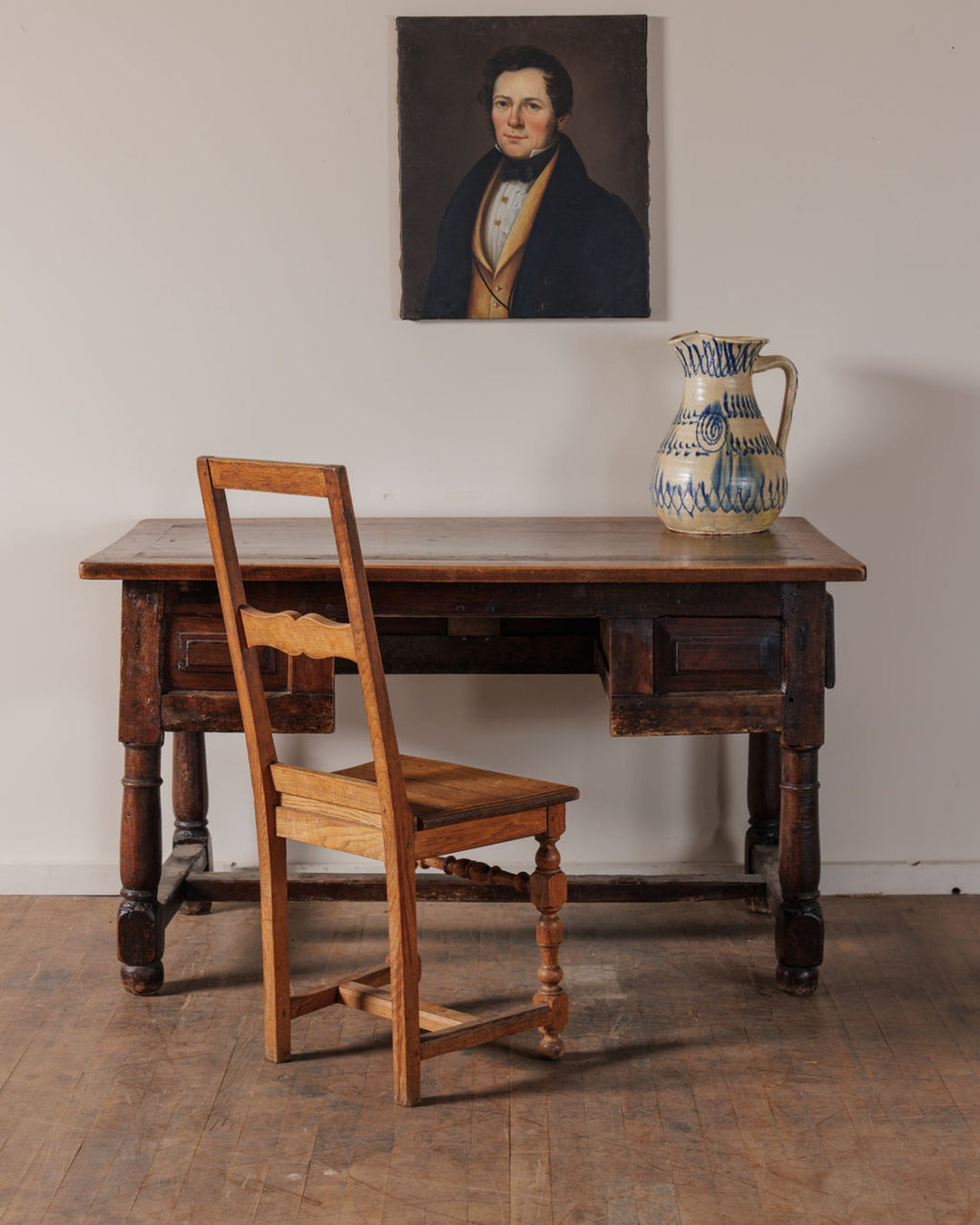 An antique wooden desk and chair, with a bust portrait on the wall and a blue and white ceramic pitcher on the desk.