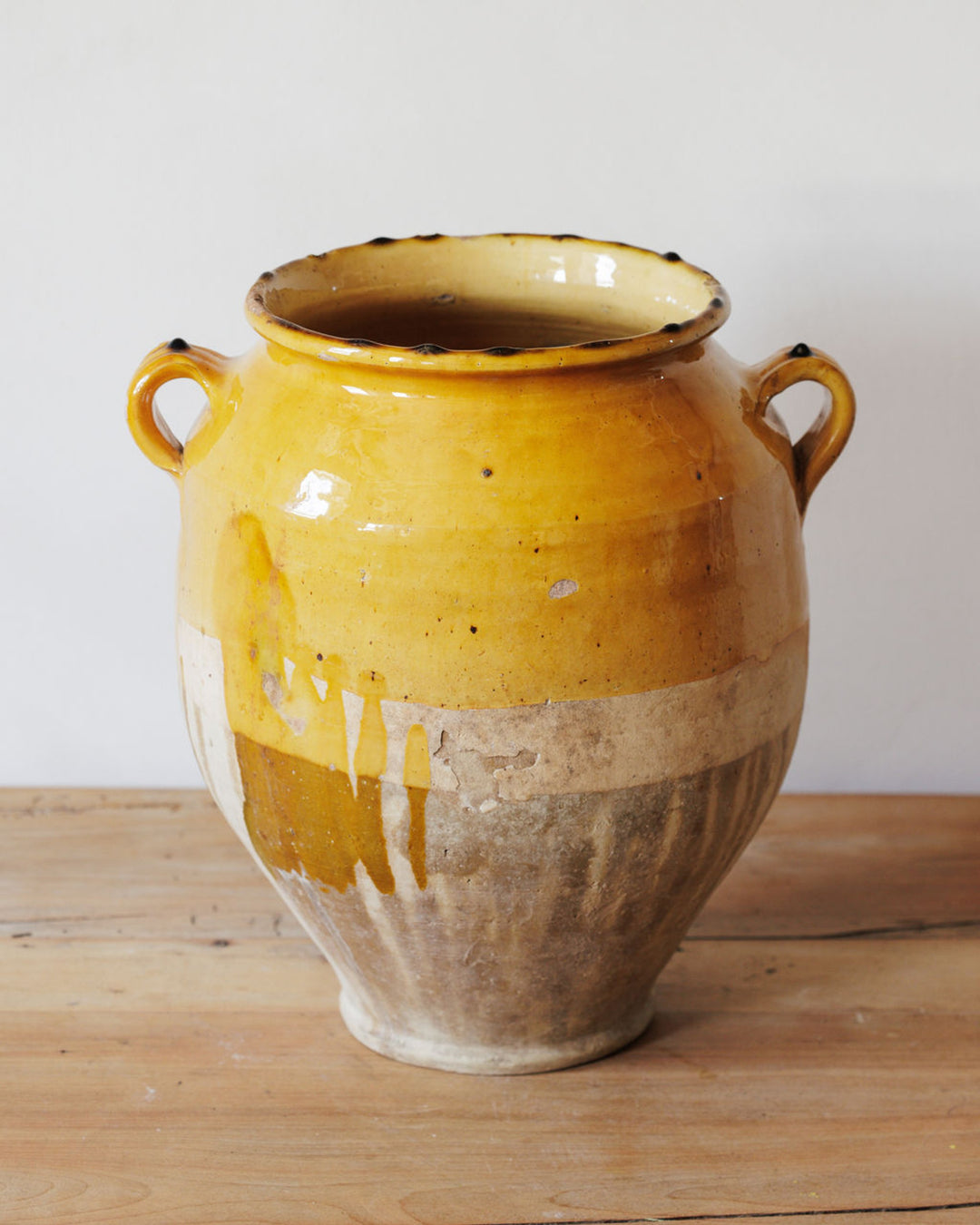 A two-handled ceramic pot with a yellow glazed top that drips over its unglazed, rustic base, sitting on a wooden surface.