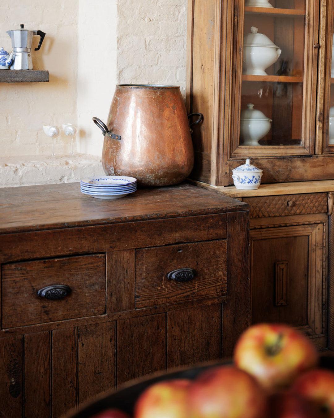A rustic kitchen with a large copper pot, antique wooden furniture, stacked plates, and apples in the foreground.