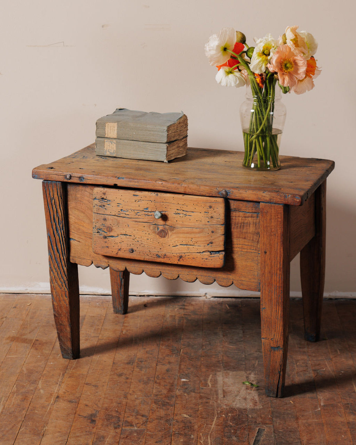 A distressed wooden cabinet with a drawer, topped by old books and a vase of white, orange, and peach poppies.