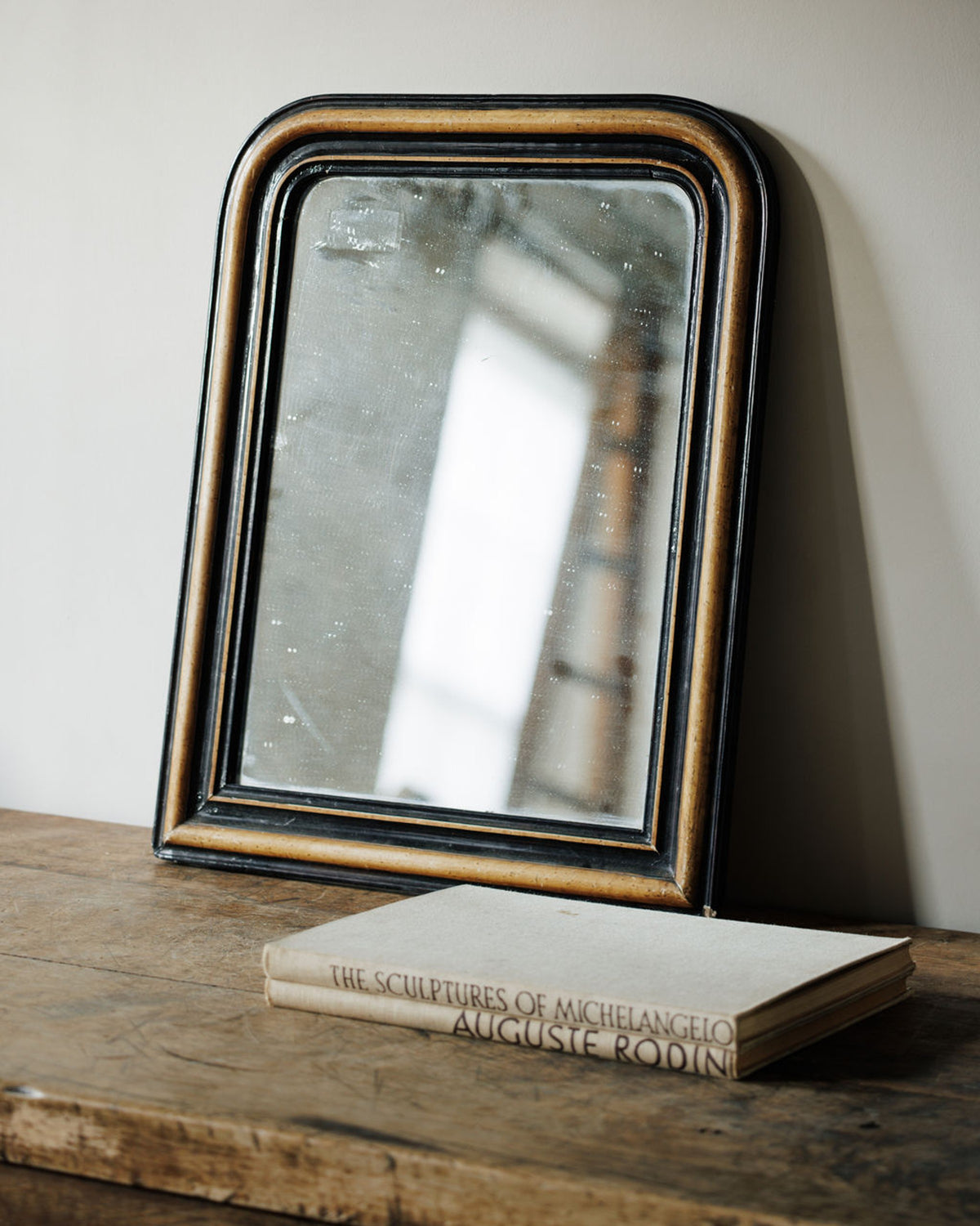 Old black and gold mirror on a rustic wooden table with 'The Sculptures of Michelangelo Auguste Rodin' book.