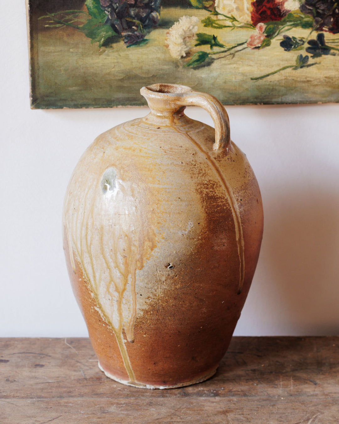 Rustic stoneware jug with a drippy, two-tone glaze on a wooden table, with a floral painting behind it.