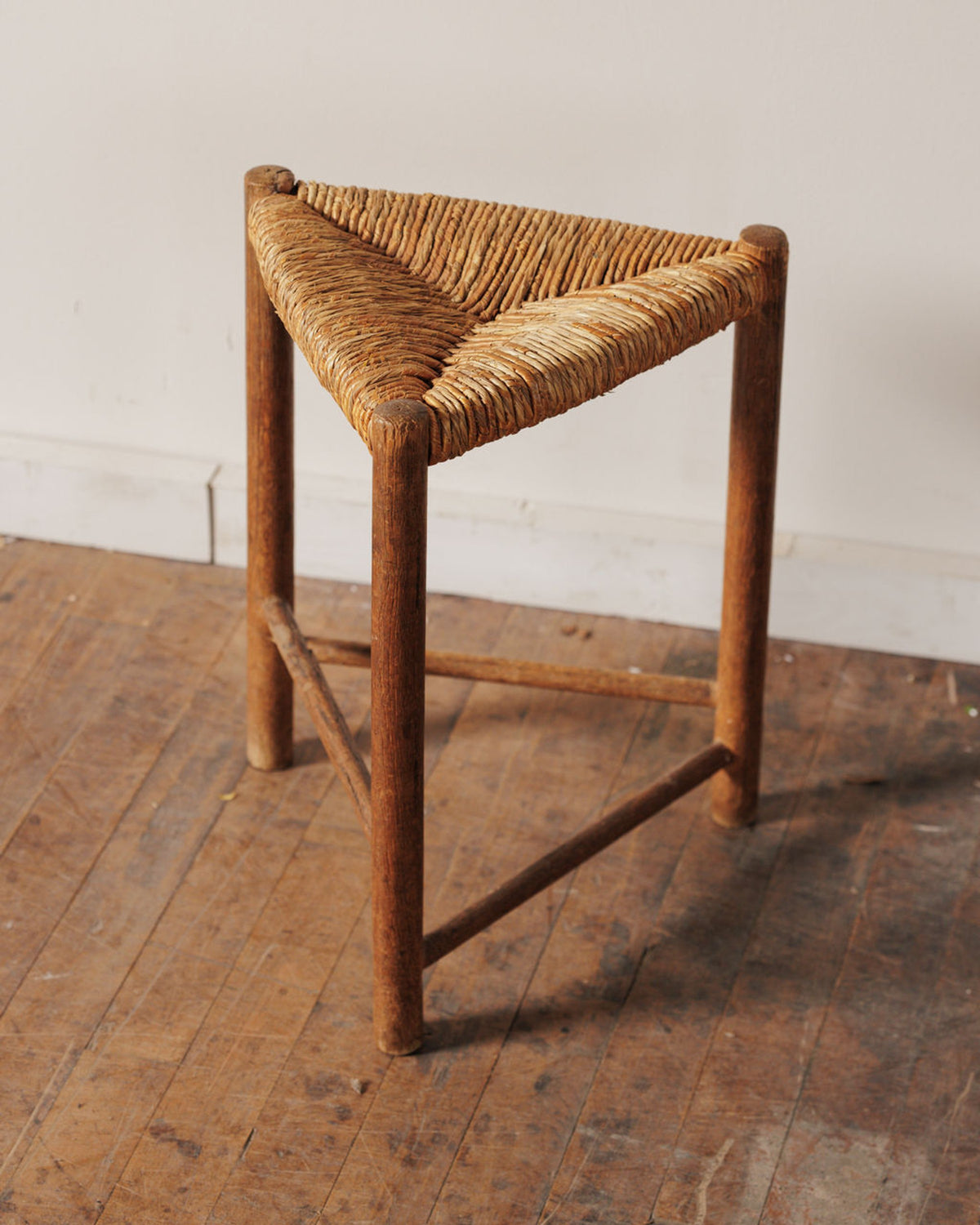 A three-legged wooden stool with a triangular woven rush seat on a wooden floor.