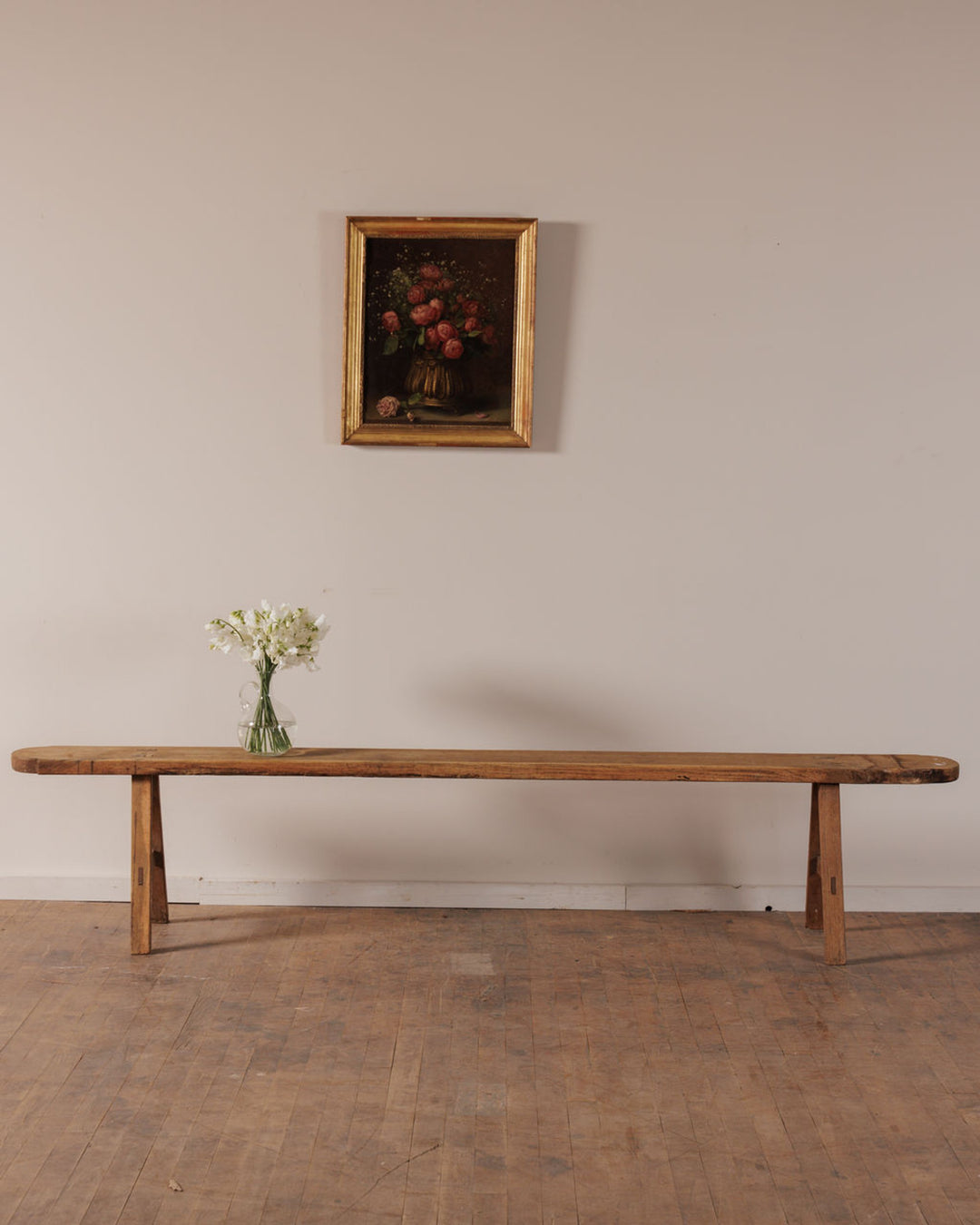 A long rustic wooden bench with a vase of white flowers, below a framed painting of red flowers.
