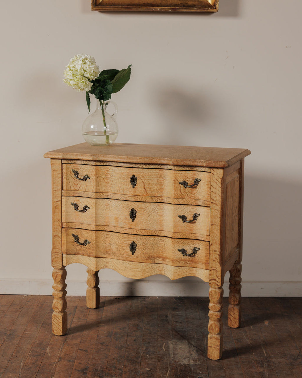 A light wood commode with three drawers and carved legs, topped with a vase holding a white hydrangea.