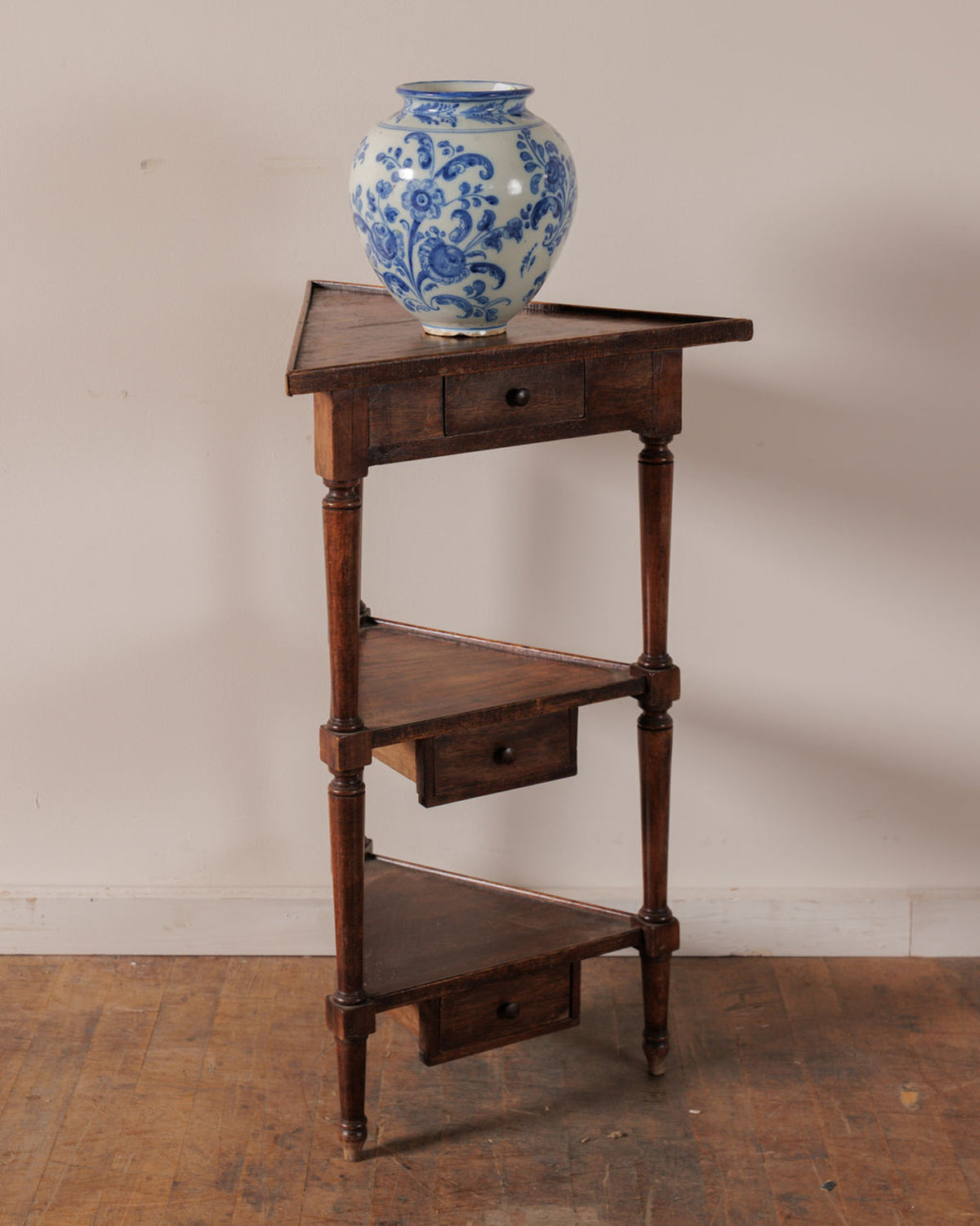 A dark wood three-tier corner unit with integrated drawers, topped by a blue and white floral vase.