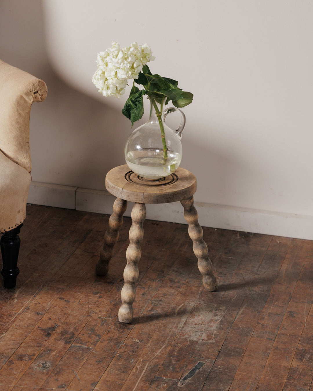 A glass pitcher of white hydrangeas on a rustic wooden stool with bobbin legs.