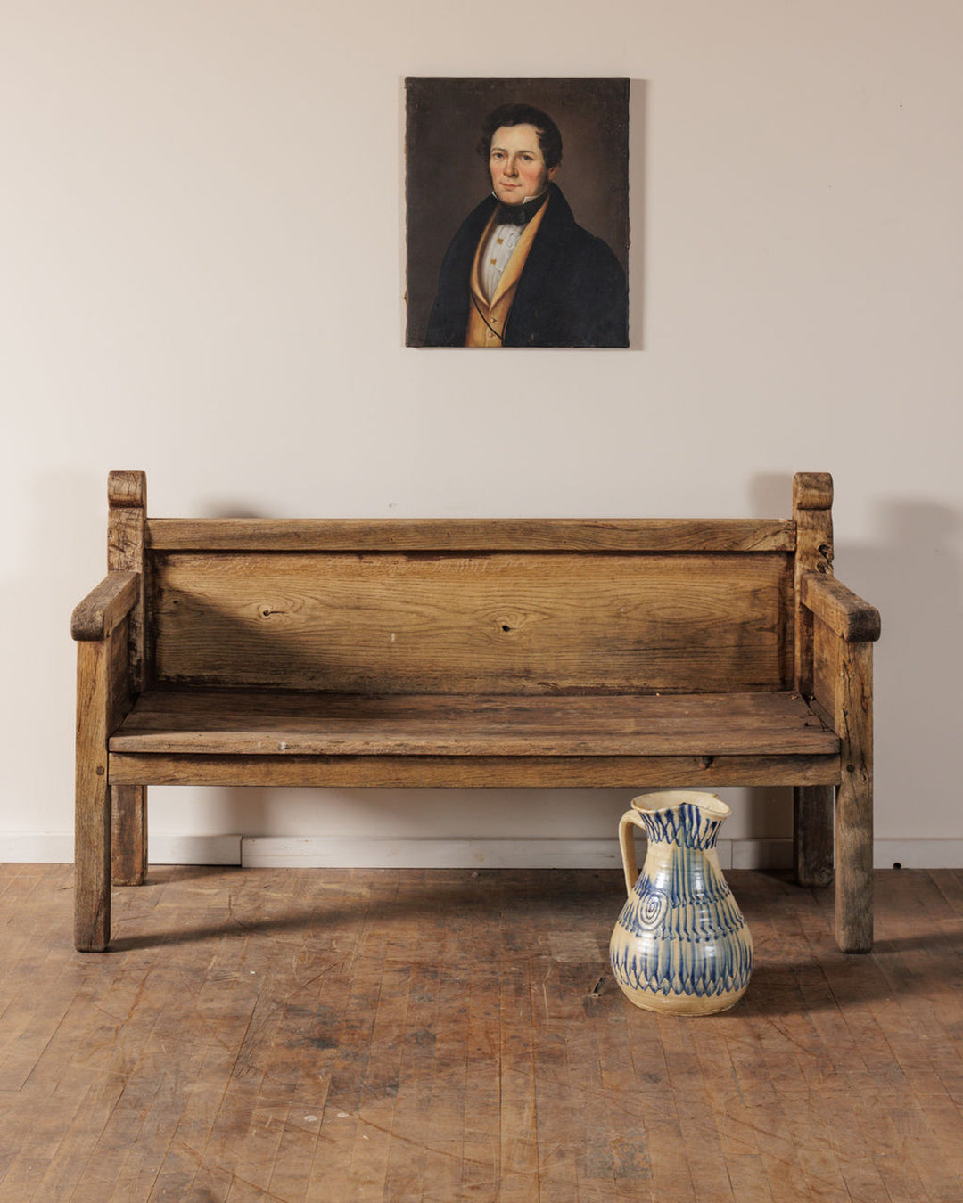 A painting of a man hangs above an antique wooden bench with a patterned ceramic pitcher on the floor beside it.