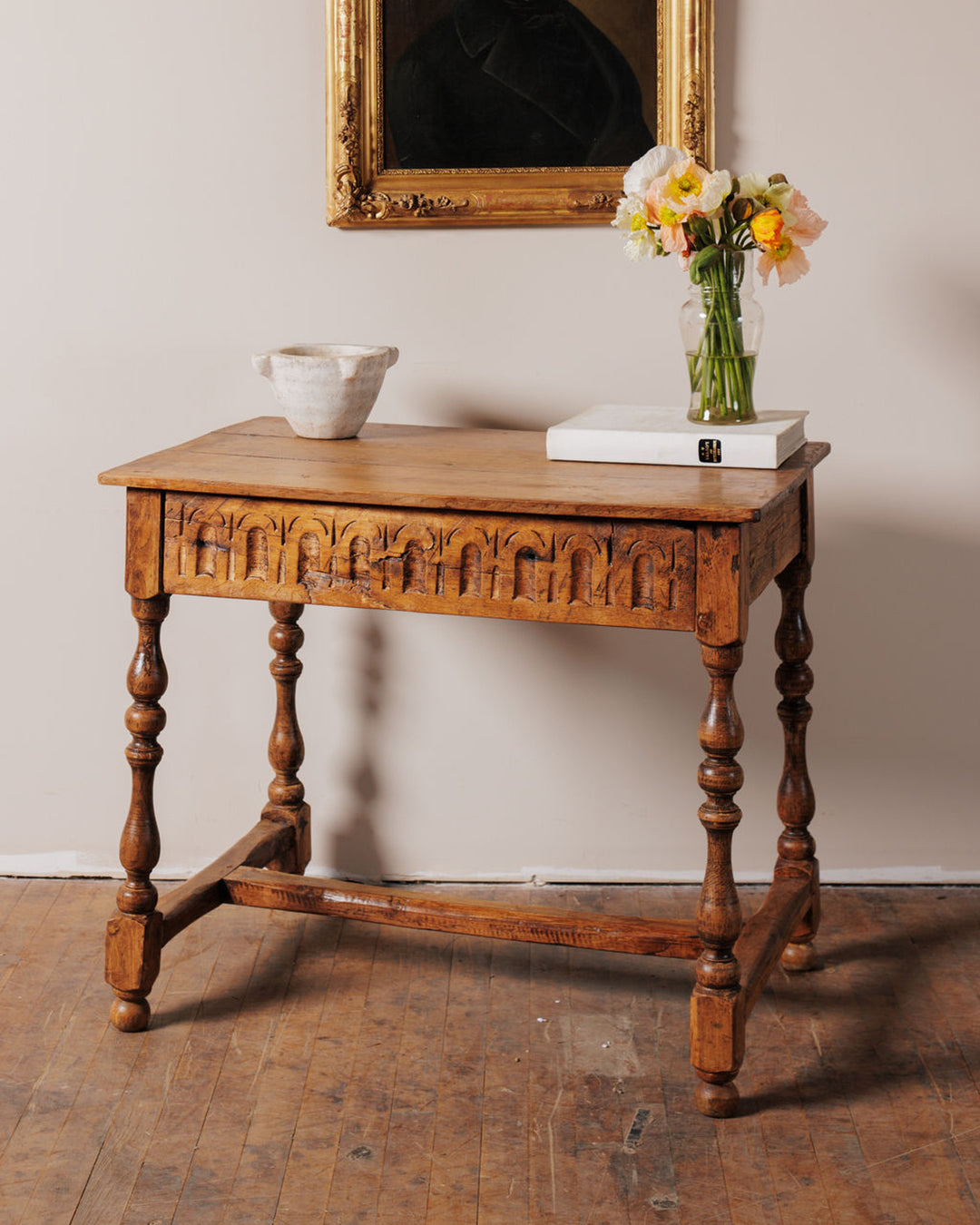 Carved wooden table with turned legs, holding a white bowl, books, and a vase of colorful poppies, below a framed portrait.