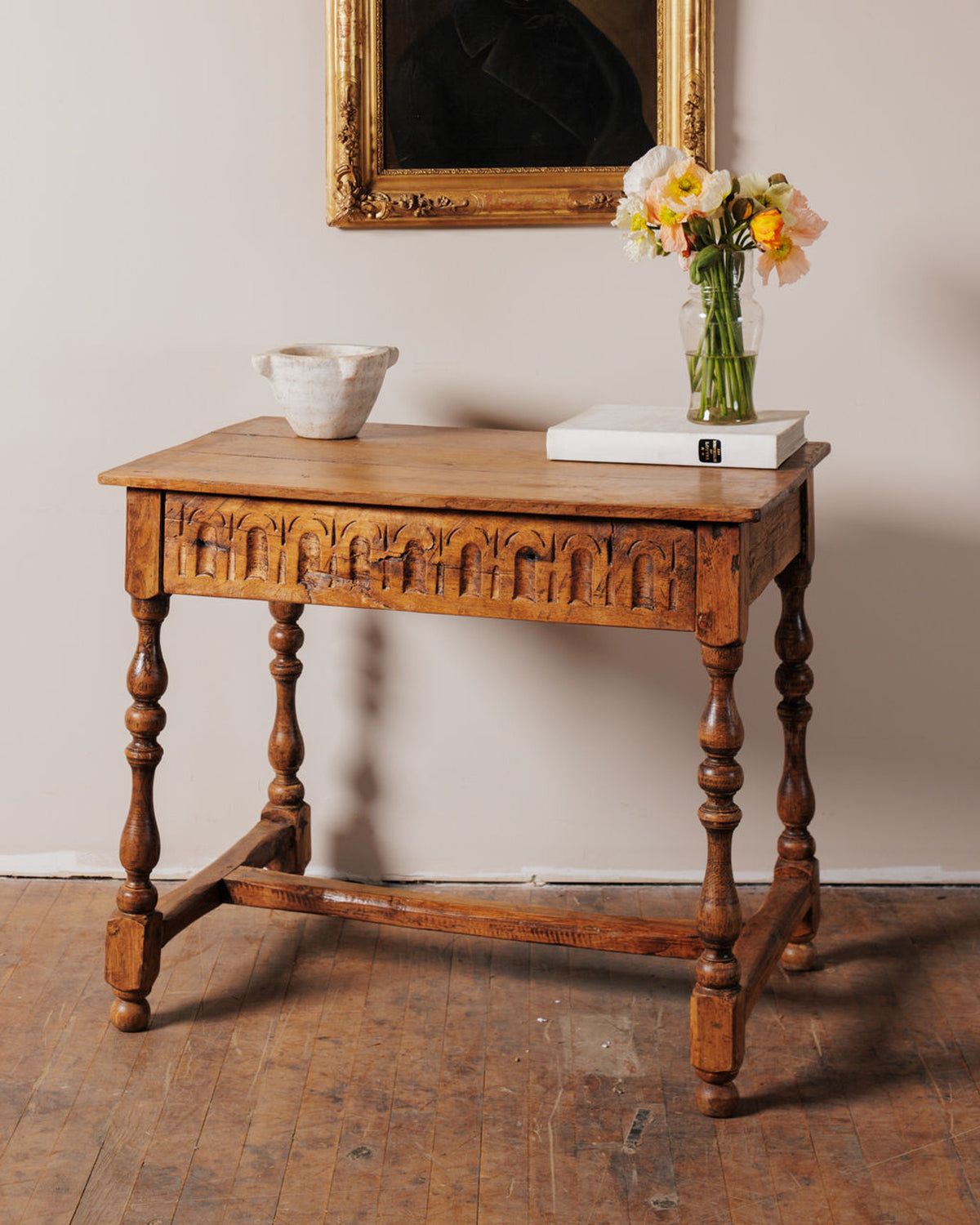 Carved wooden table with turned legs, holding a white bowl, books, and a vase of colorful poppies, below a framed portrait.
