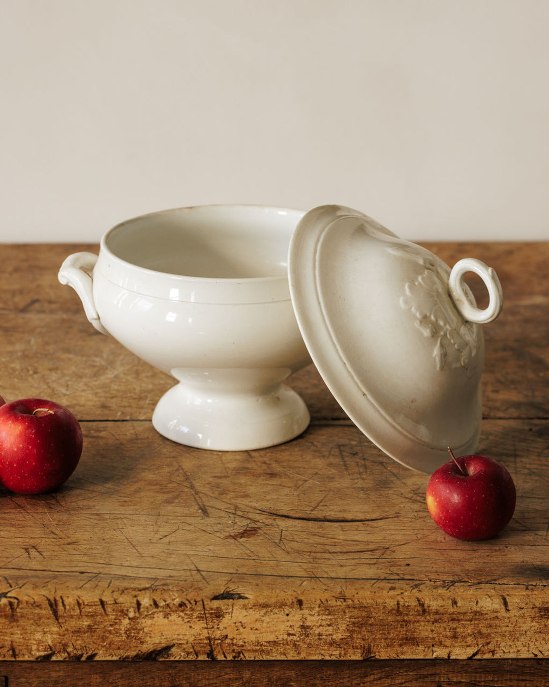 White ceramic tureen with tilted lid and two red apples on a rustic wooden table.