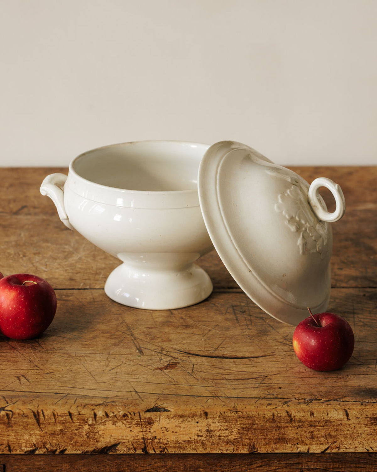 White ceramic tureen with tilted lid and two red apples on a rustic wooden table.