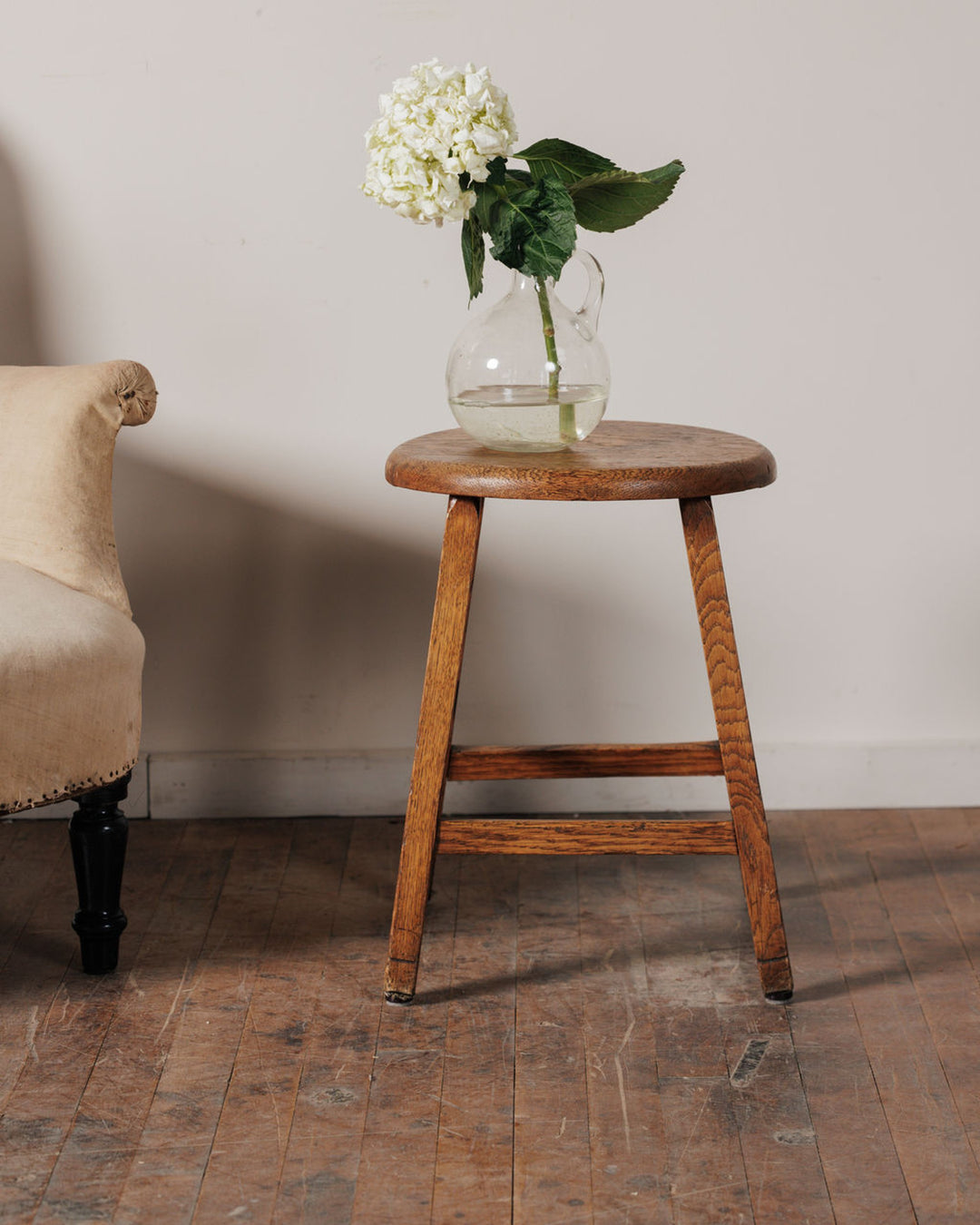 White hydrangea in a glass pitcher on a round wooden stool, with a beige armchair and wood floor.