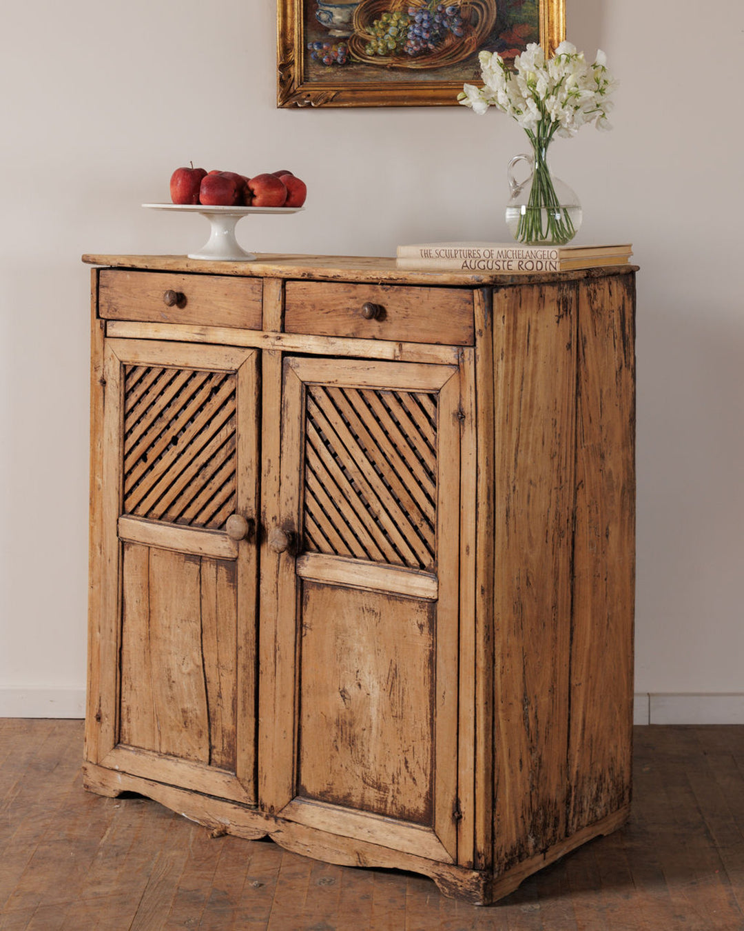 Rustic light wood cabinet with two drawers and two doors featuring diagonal slat details, topped with apples, white flowers, and a book.