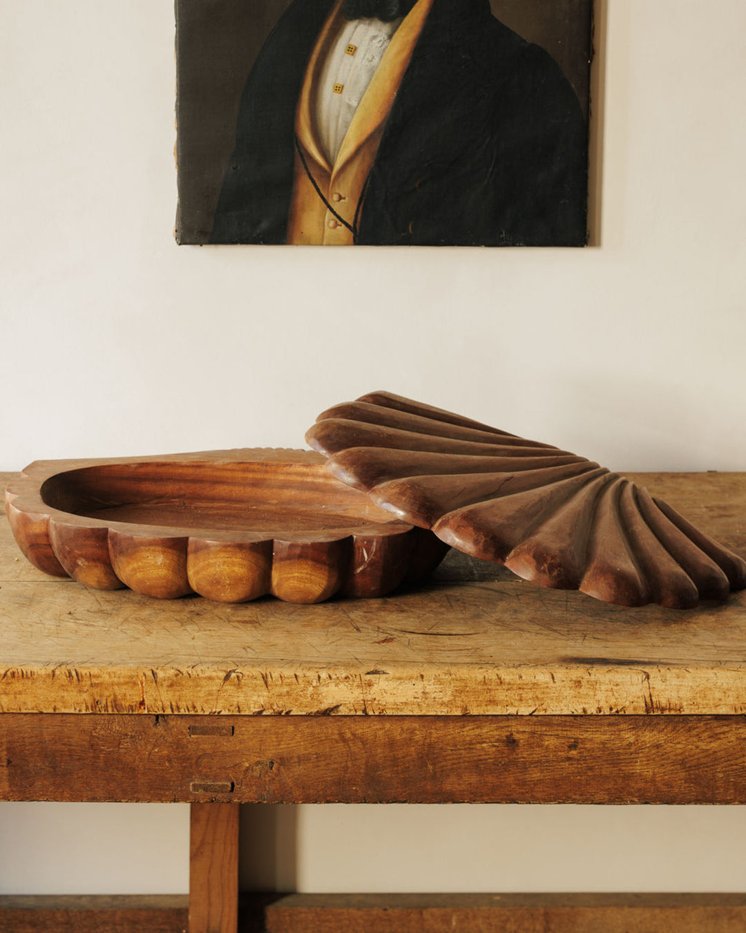 A carved wooden shell-shaped dish with its lid ajar, sitting on a rustic wooden table, with a portrait painting in the background.