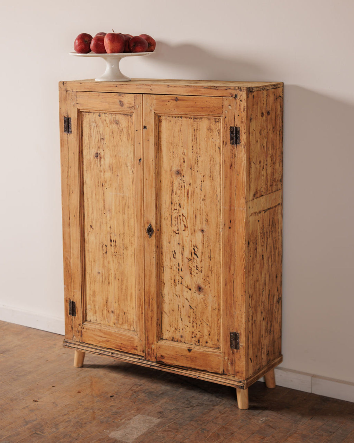 Distressed wooden cabinet with two doors, topped with a white pedestal bowl of red apples.