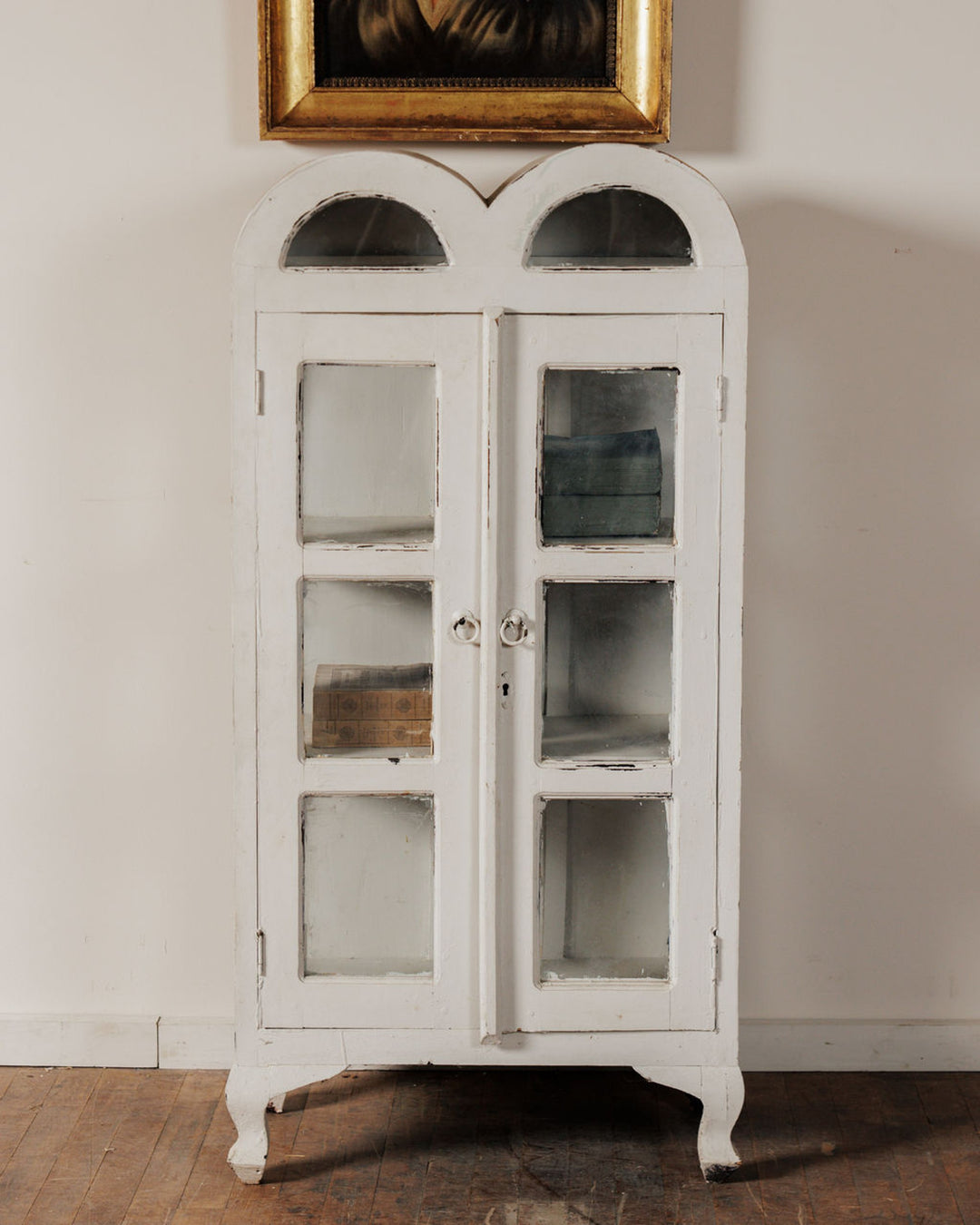 A distressed white wooden display cabinet with glass doors and a scalloped top, standing on a wooden floor below a framed painting.