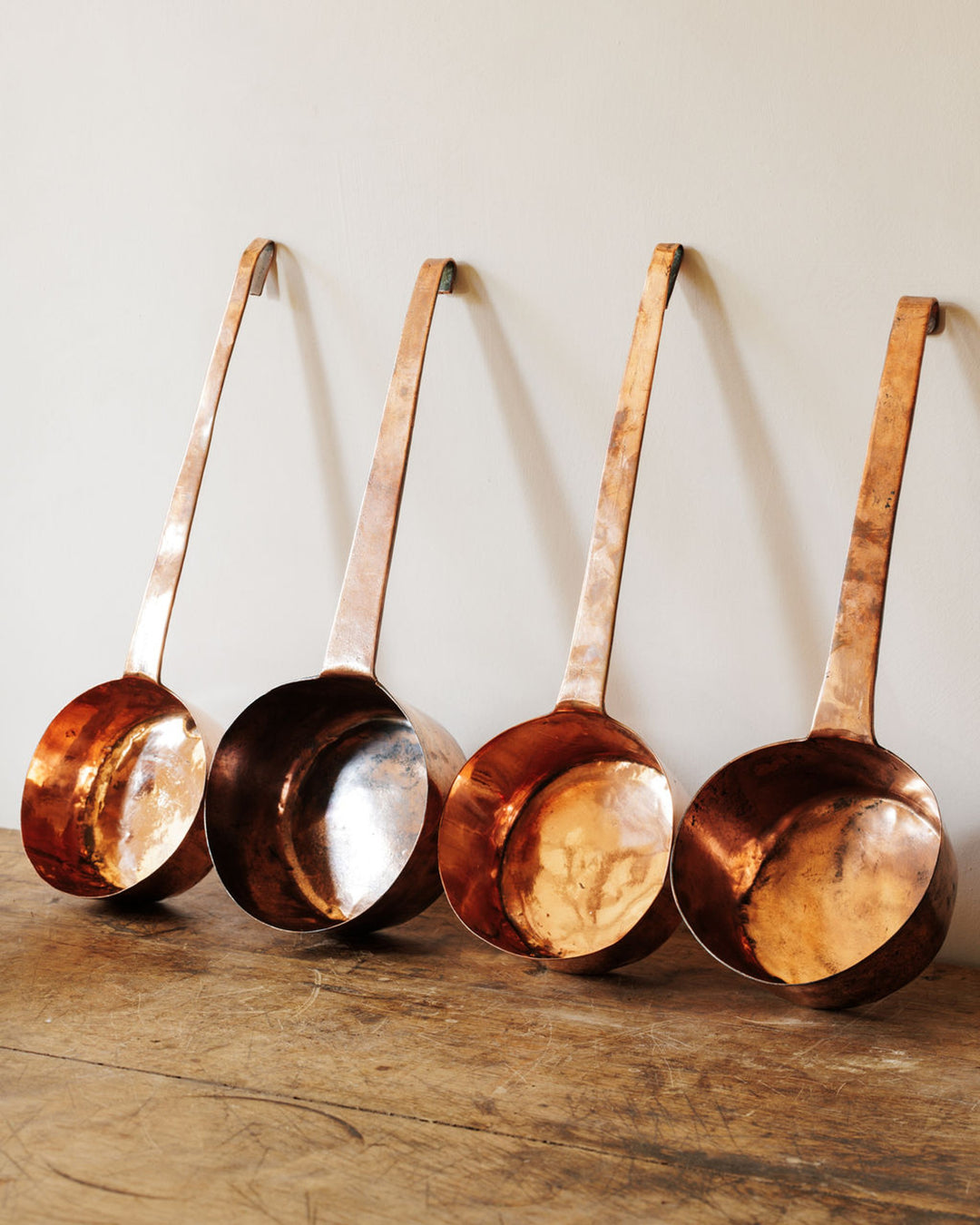 Four copper ladles hanging against a light wall, resting on a wooden surface.