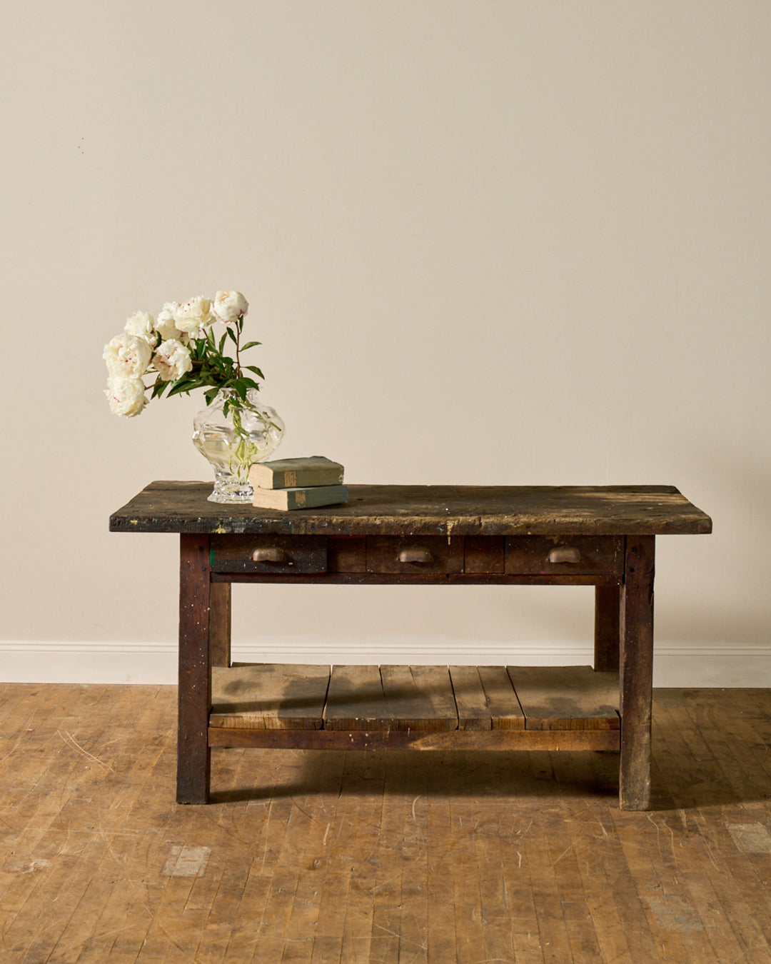 A rustic wooden table holding a vase of white flowers and two old books, against a light wall and wooden floor.