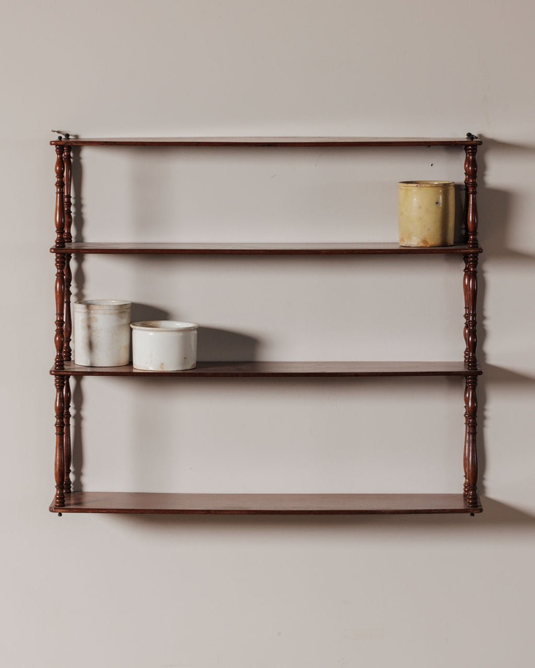 A four-tier wooden wall shelf with turned supports, holding one yellow and two white ceramic crocks.