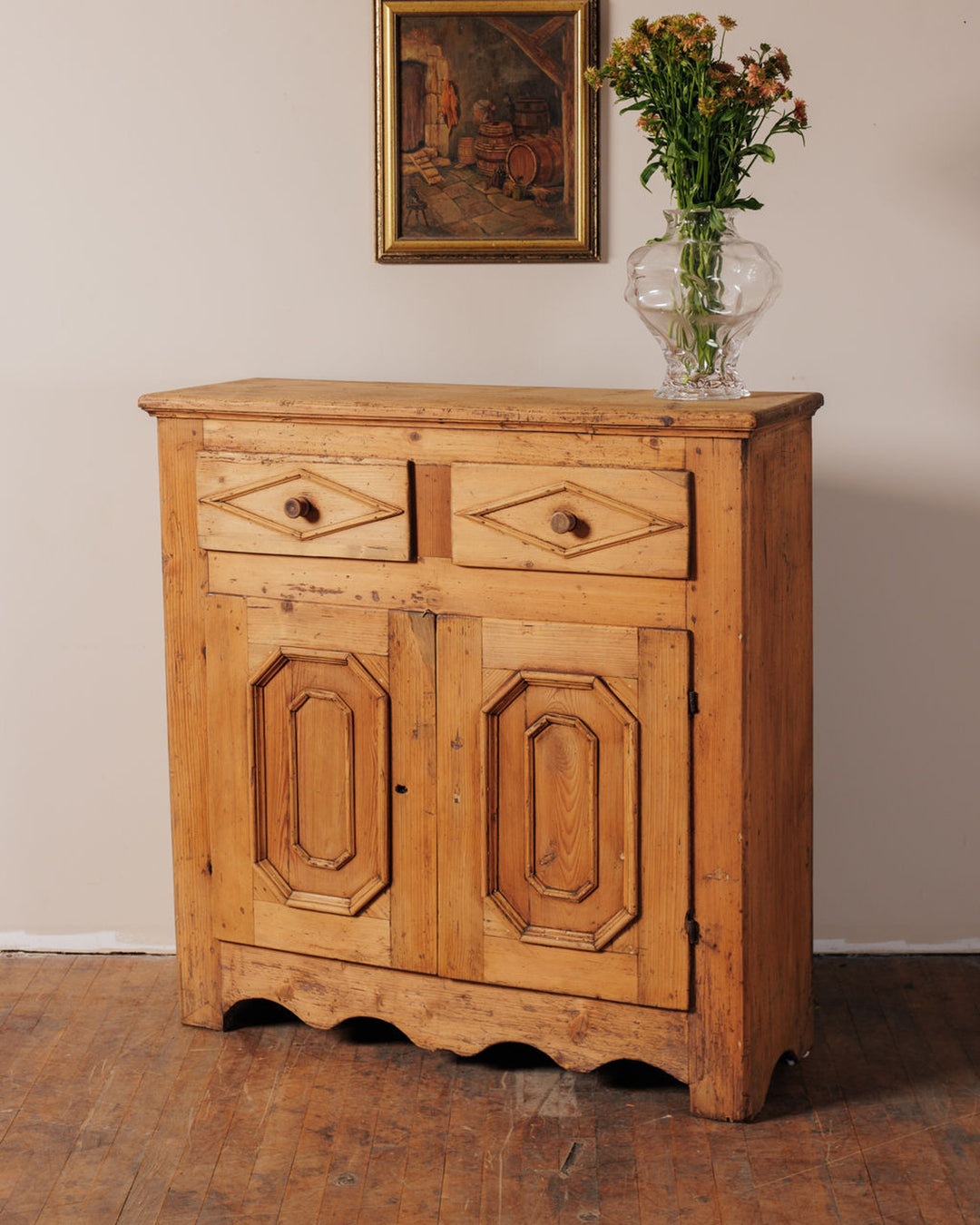 Antique pine cabinet with diamond-carved drawers, paneled doors, and a scalloped base, topped with a vase of flowers, with a framed painting above.