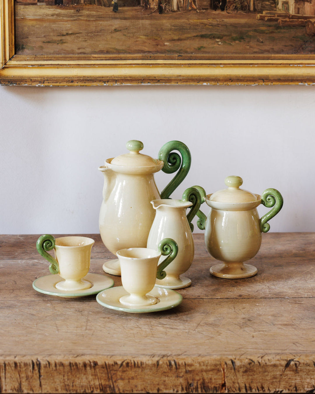 Cream-colored ceramic tea set with green scroll handles on a wooden table, below a framed painting.