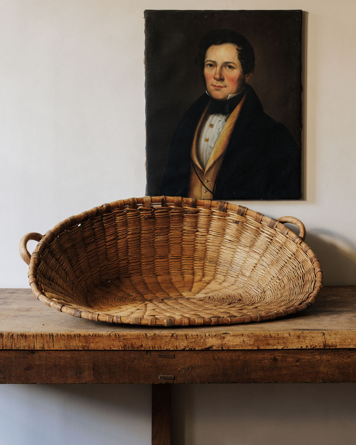 An oval woven basket sits on a rustic wooden table, with a portrait of a man in formal attire hanging on the wall behind it.