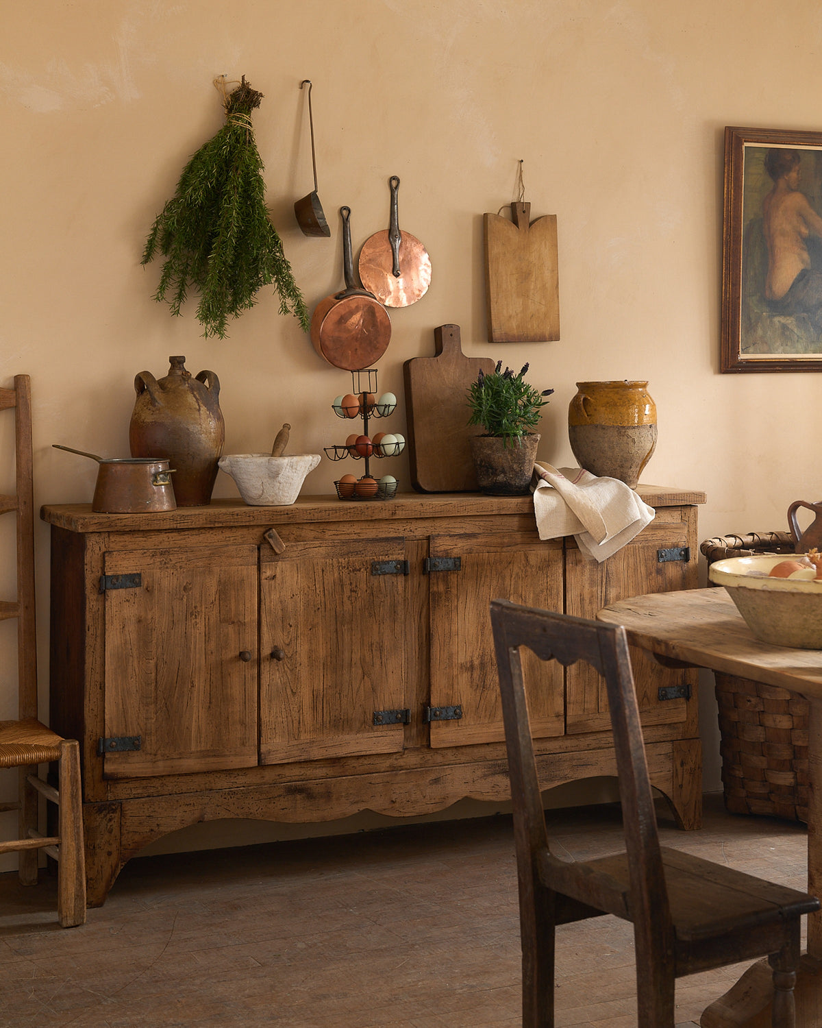 A rustic kitchen featuring a wooden sideboard, hanging copper pots, herbs, and pottery on a beige wall.