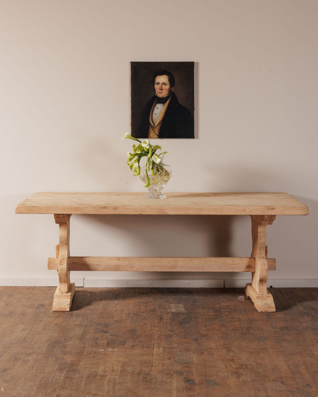 A rustic wooden table with a vase of white calla lilies, beneath a portrait of a man, against a light wall and wooden floor.