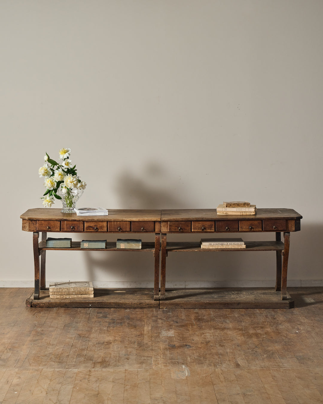 A long, rustic wooden console table with multiple drawers and shelves, decorated with books and a vase of white flowers.