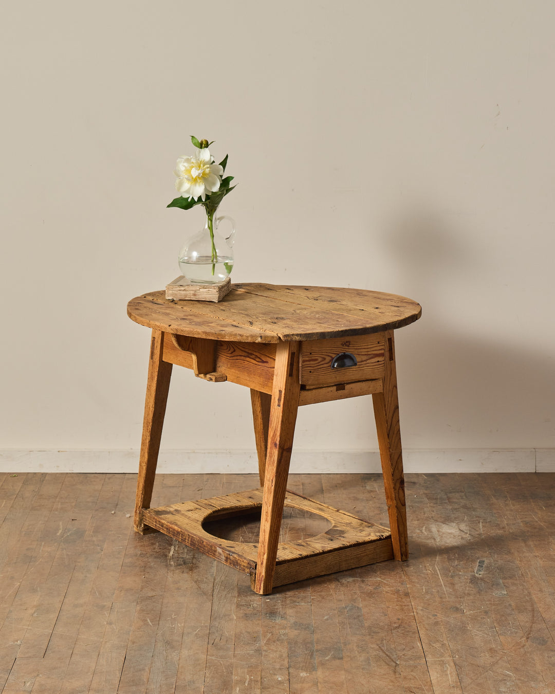 Rustic pine side table with an oval top, a drawer, and a lower shelf with a circular cutout, holding a white peony in a glass pitcher.