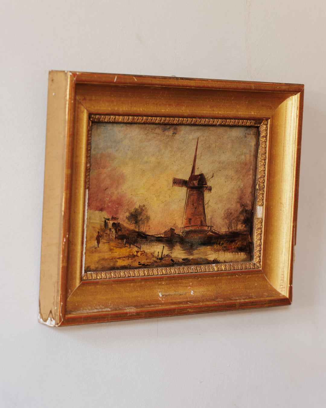 A landscape painting of a windmill by water in a worn gold frame.