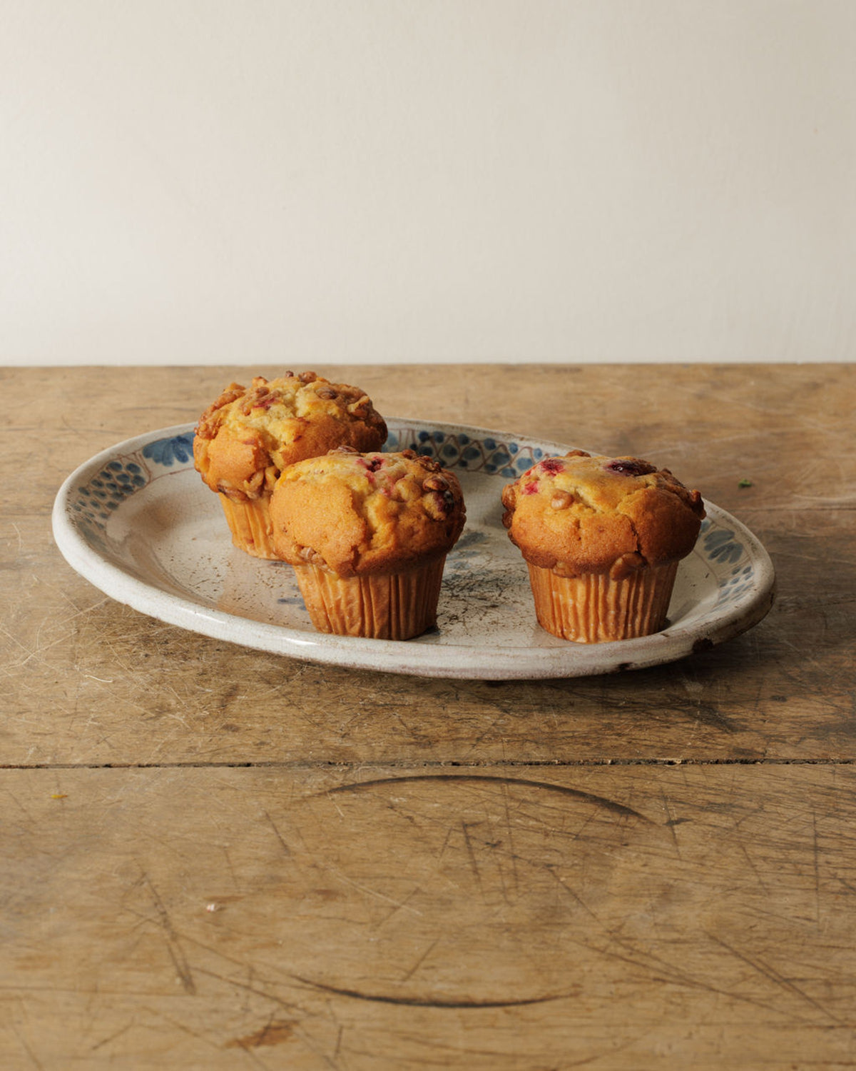 Three golden muffins with berry and nut toppings on a blue and white patterned oval plate, on a rustic wooden table.