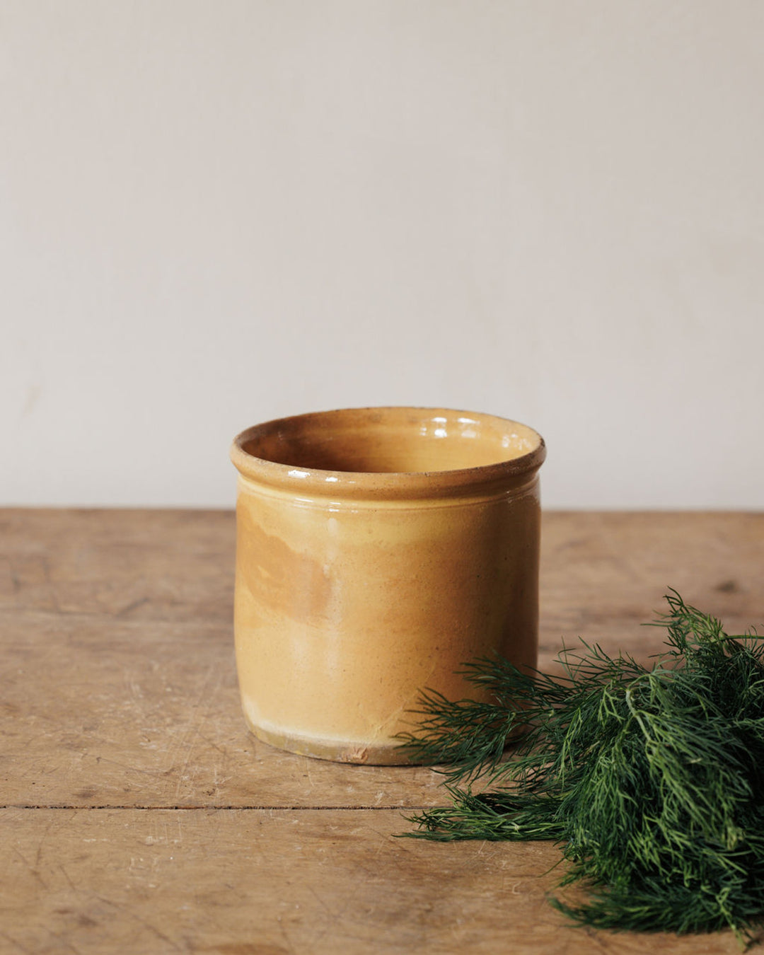 A mustard-colored ceramic jar sits on a wooden table with a bunch of fresh dill to its right.