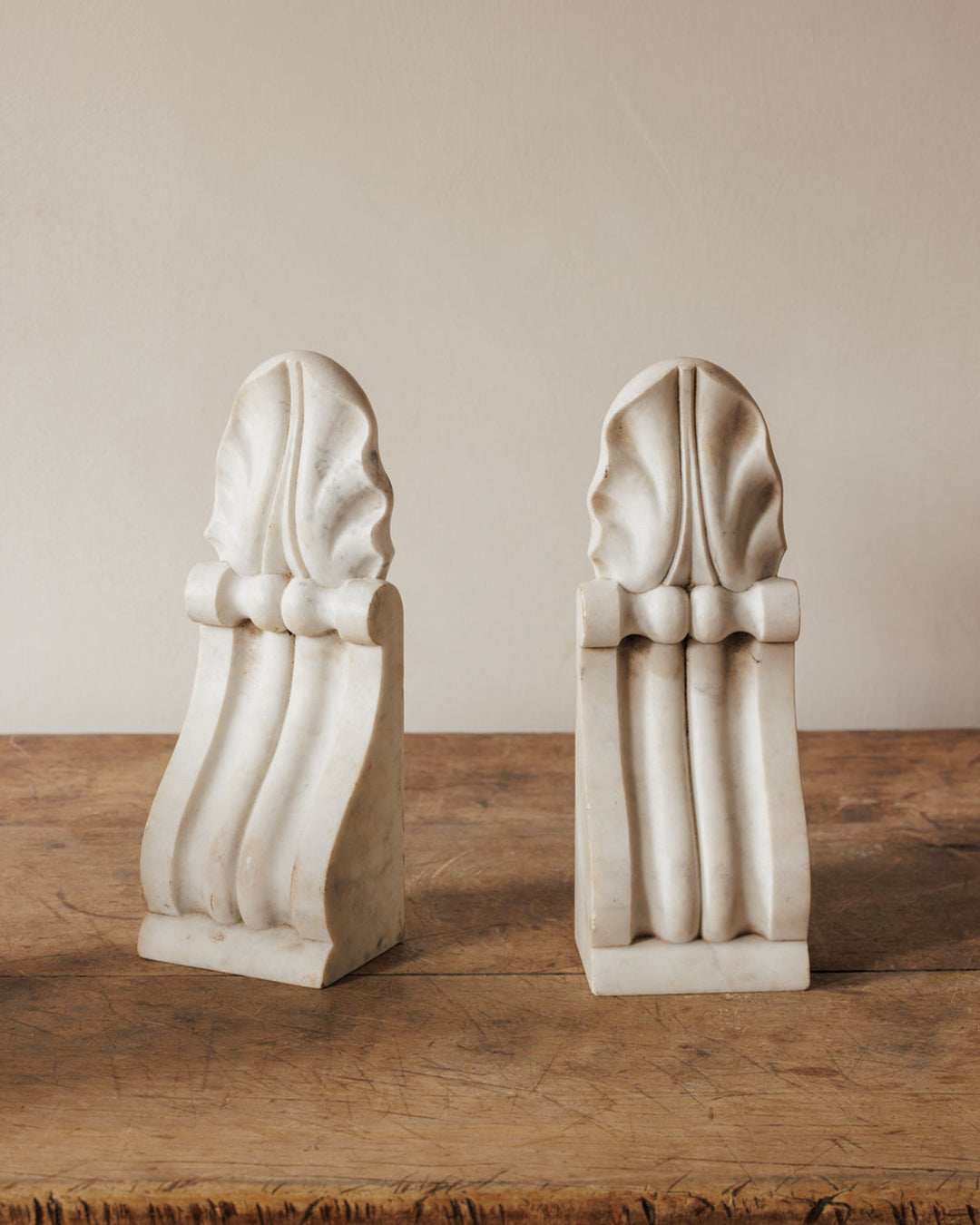 A pair of white marble architectural elements with carved leaf motifs and fluted supports on a wooden table.