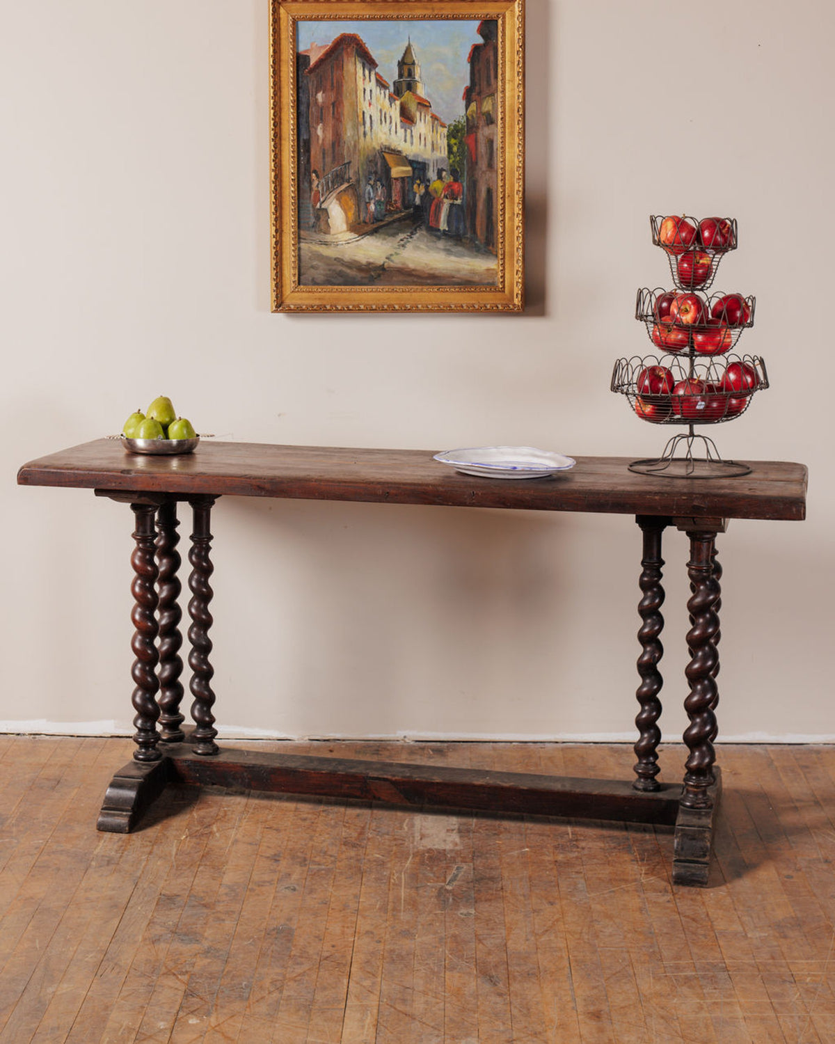 A dark wooden console table with spiral legs holds green pears, a plate, and red apples in a tiered basket, below a framed street scene painting.