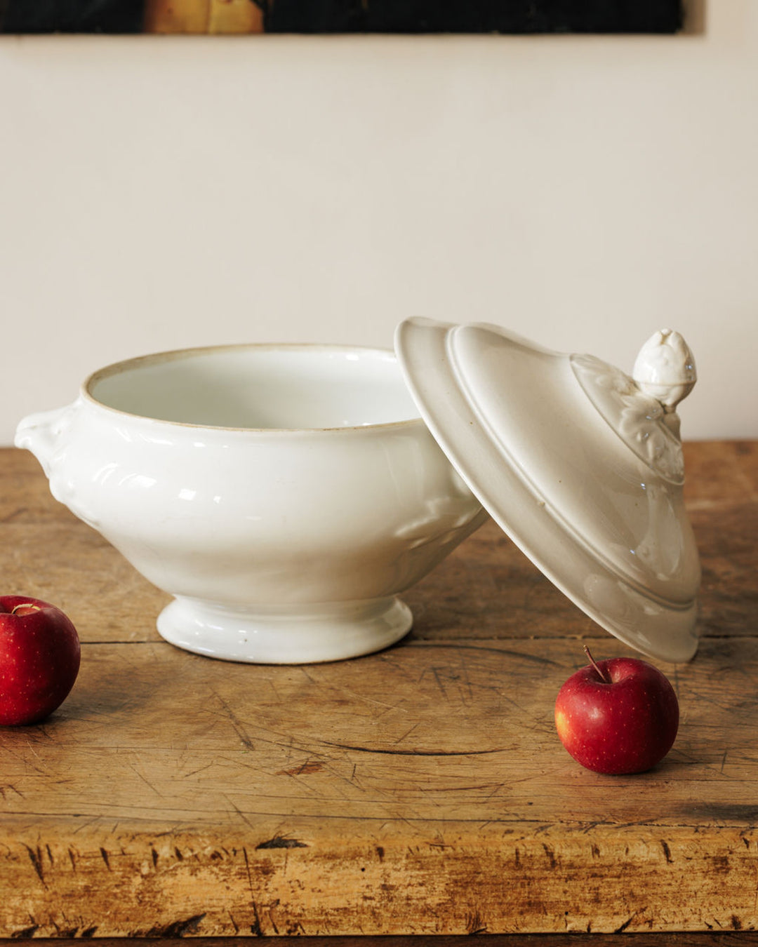 A white tureen with its lid resting beside it on a rustic wooden table, flanked by two red apples.