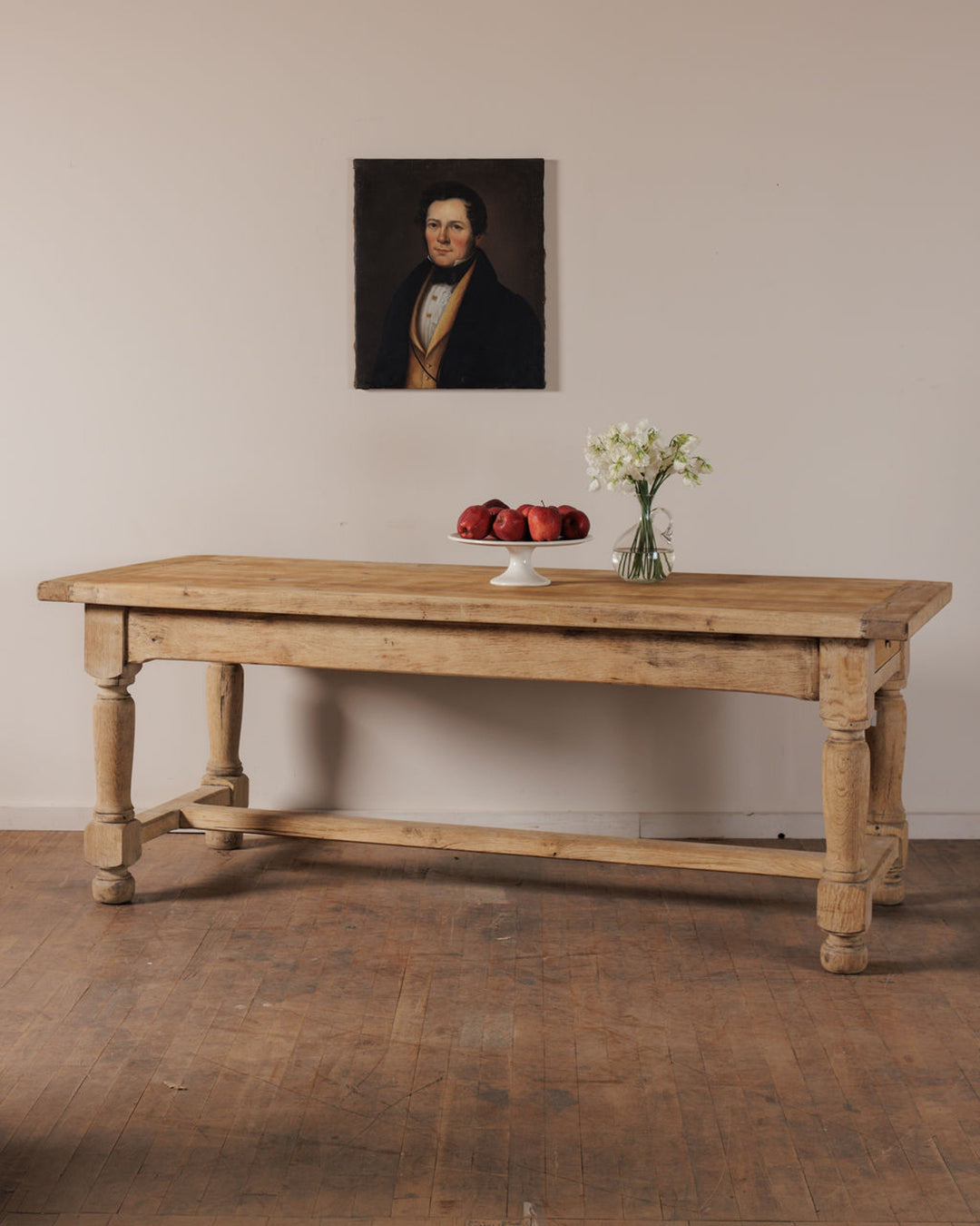 A rustic wooden table with a bowl of red apples and white flowers, beneath a portrait painting of a man.