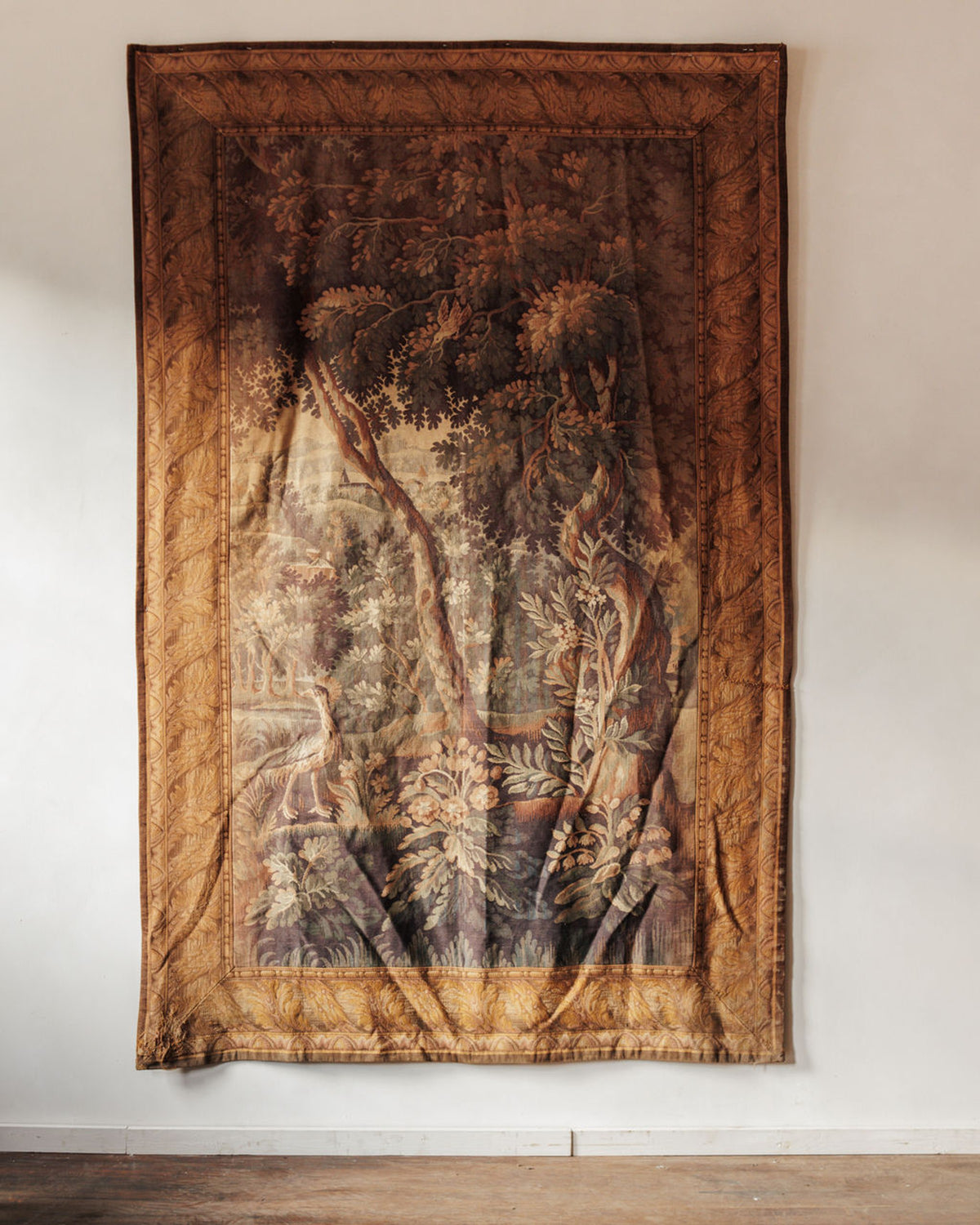 A large, antique tapestry depicting a dense forest scene with trees, foliage, and a bird, framed by an ornate border, on a white wall.