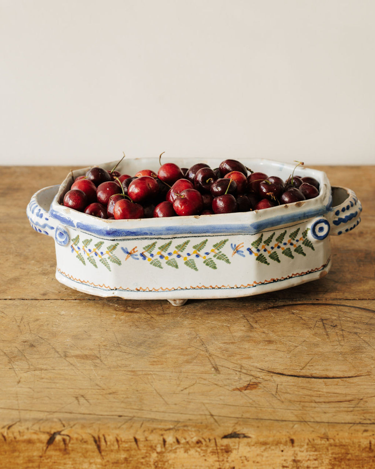A white ceramic bowl with blue and green patterns holds fresh red cherries on a wooden table.