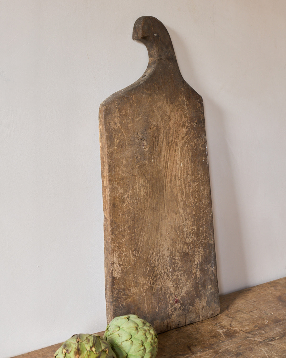Long rustic wooden cutting board leaning against a white wall, with two green tropical fruits on a wooden surface.