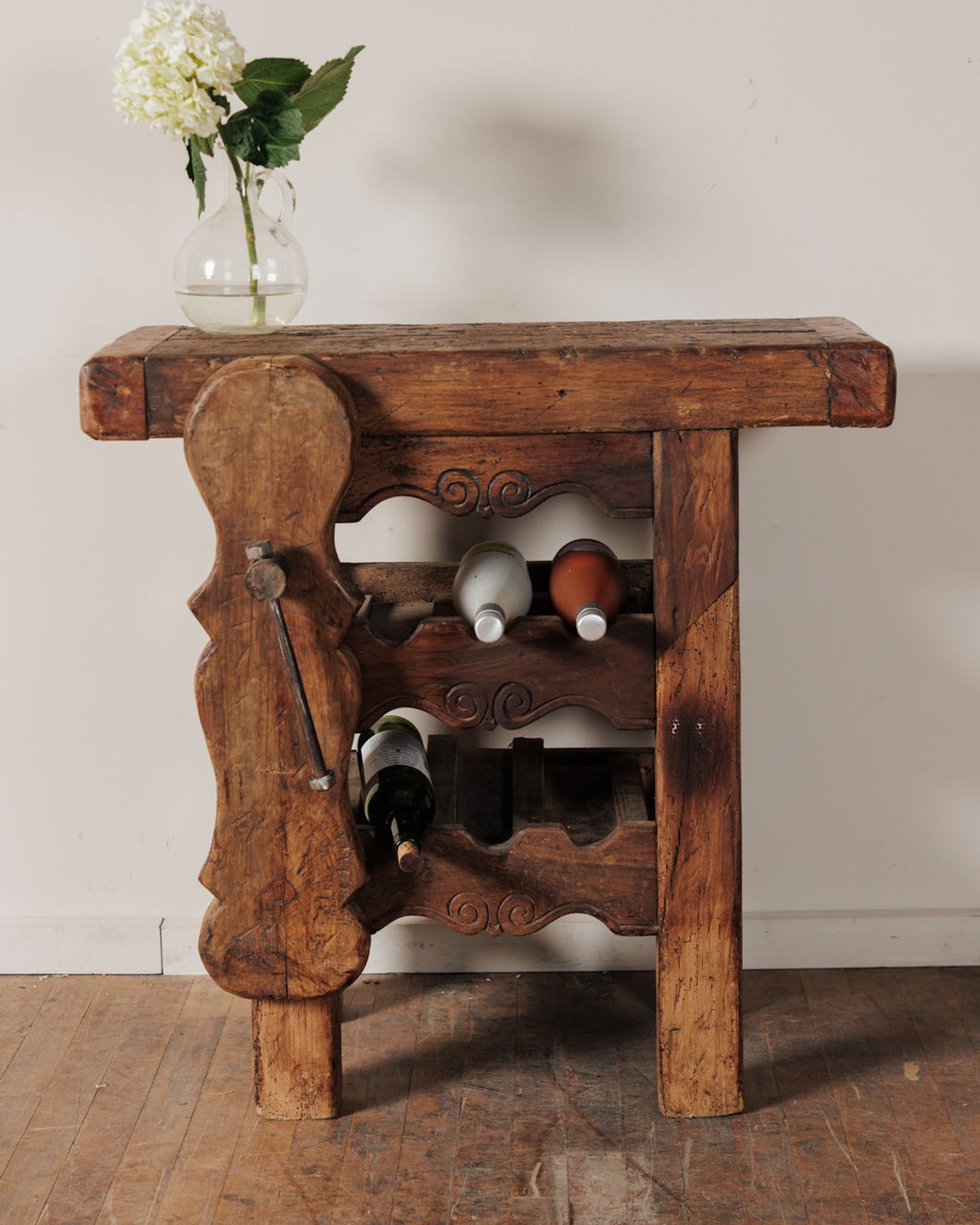 A rustic, dark wood wine rack table with carved details, holding a vase of white flowers and three wine bottles.