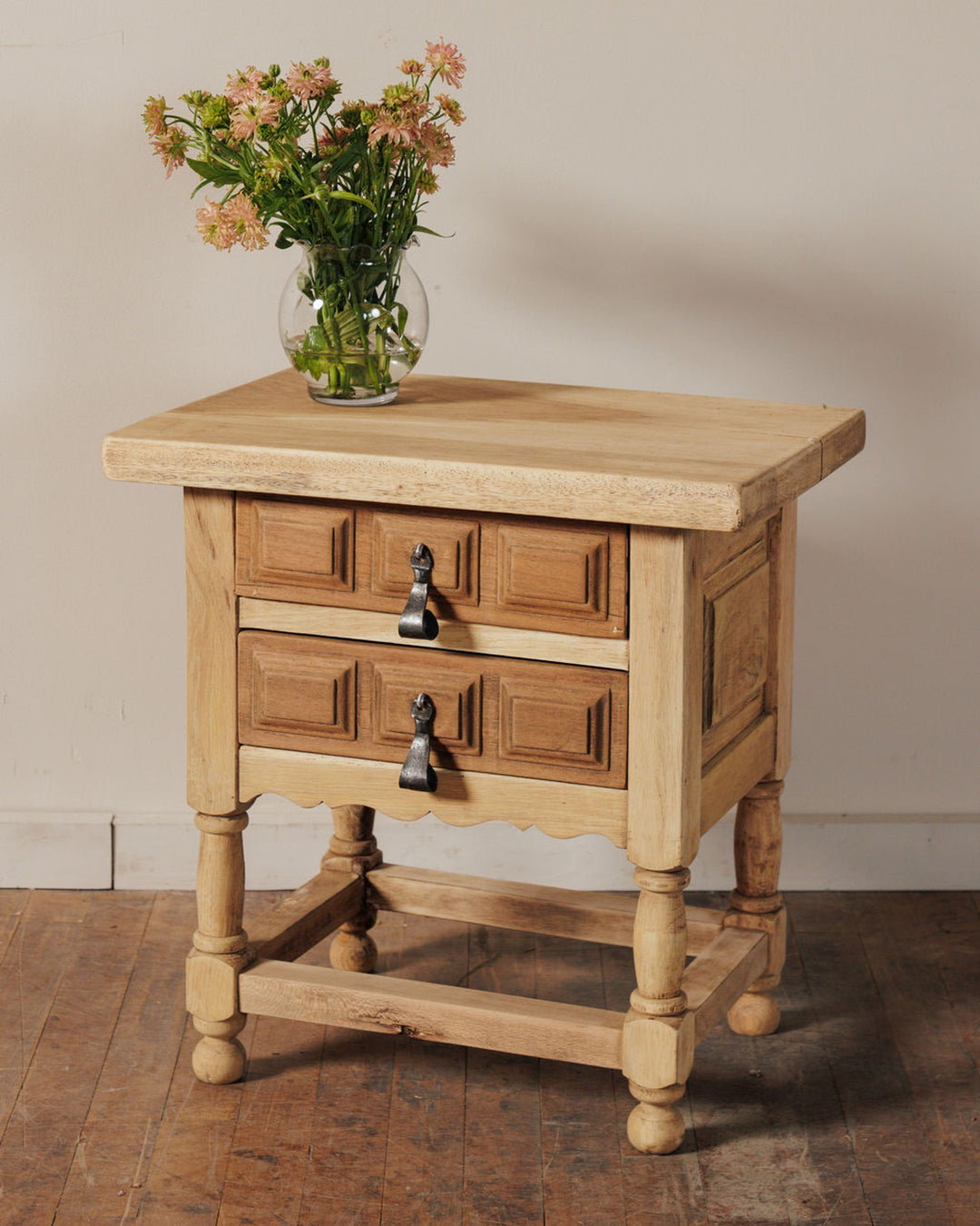 A light wood nightstand with two paneled drawers, dark metal pull handles, and a vase of pink and green flowers on top.