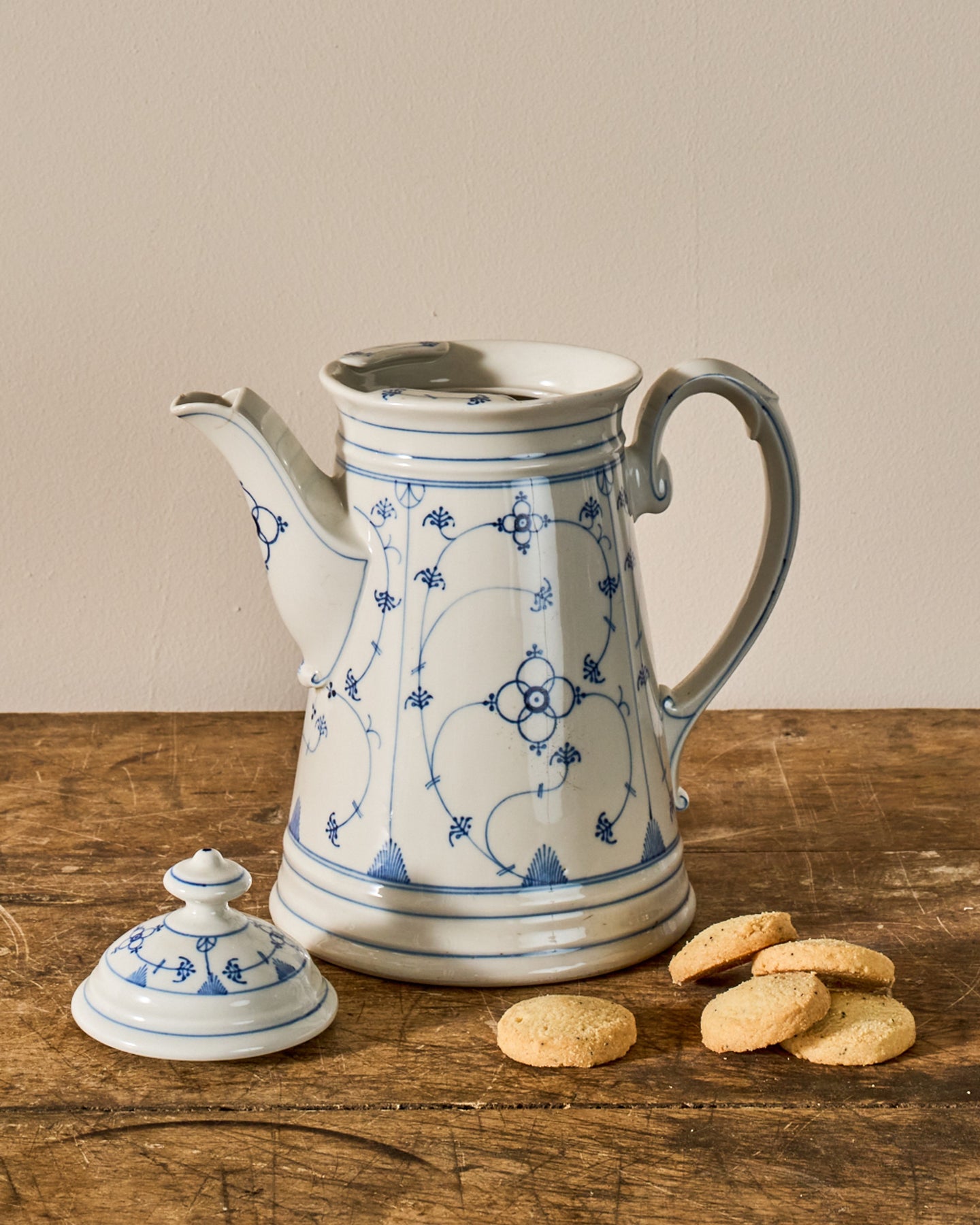 A white porcelain coffee pot with blue floral patterns and its lid off, sitting on a rustic wooden table next to several cookies.