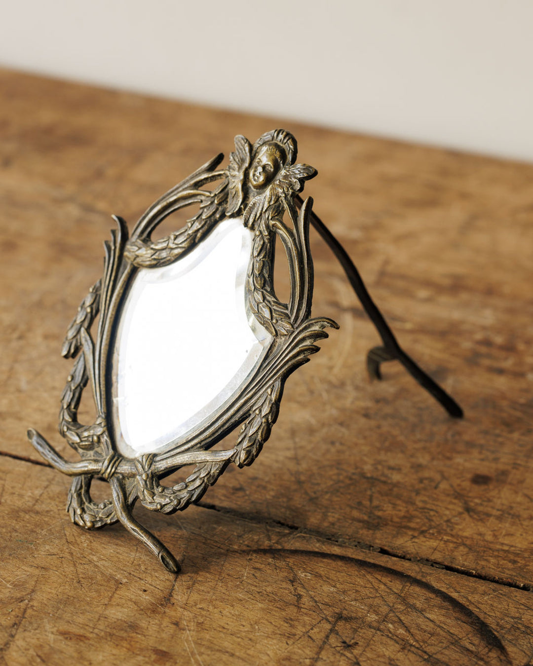 Small ornate brass vanity mirror with a cherub and laurel details on a wooden surface.