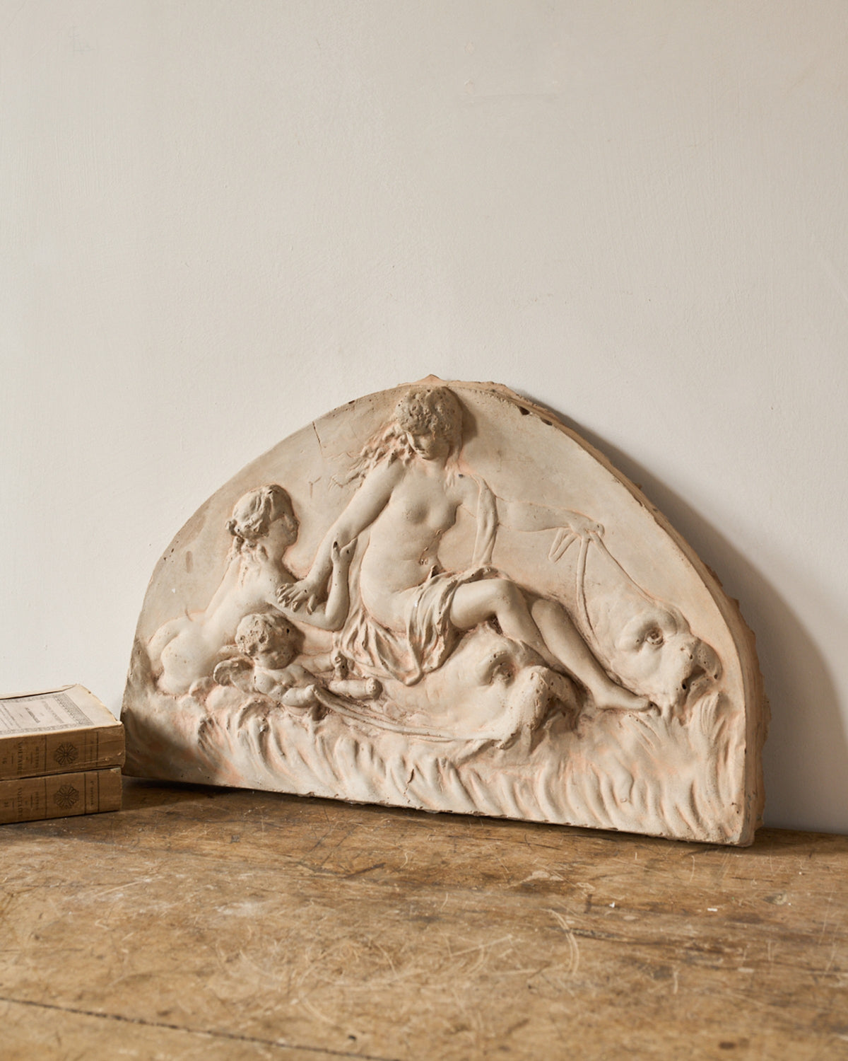A weathered demilune plaster relief sculpture depicting a reclining nude woman, putti, and a dolphin.