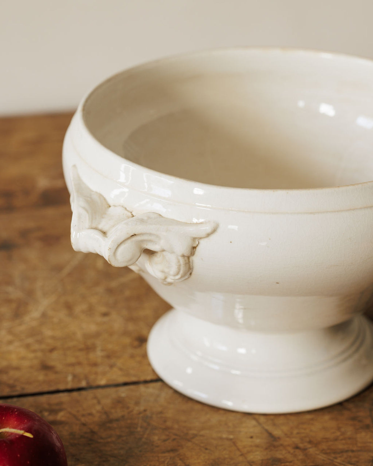 A creamy white ceramic footed bowl with ornate handles sits on a wooden table, with a red apple partially visible in the foreground.