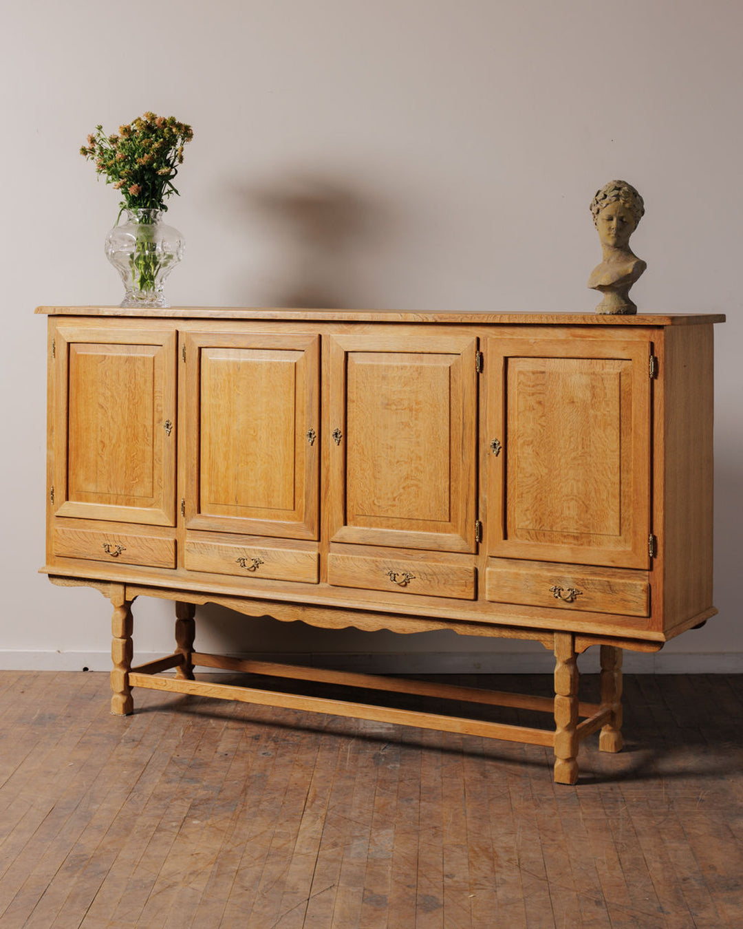 Light wood buffet with four panel doors and four drawers, adorned with a vase of flowers and a classical bust.